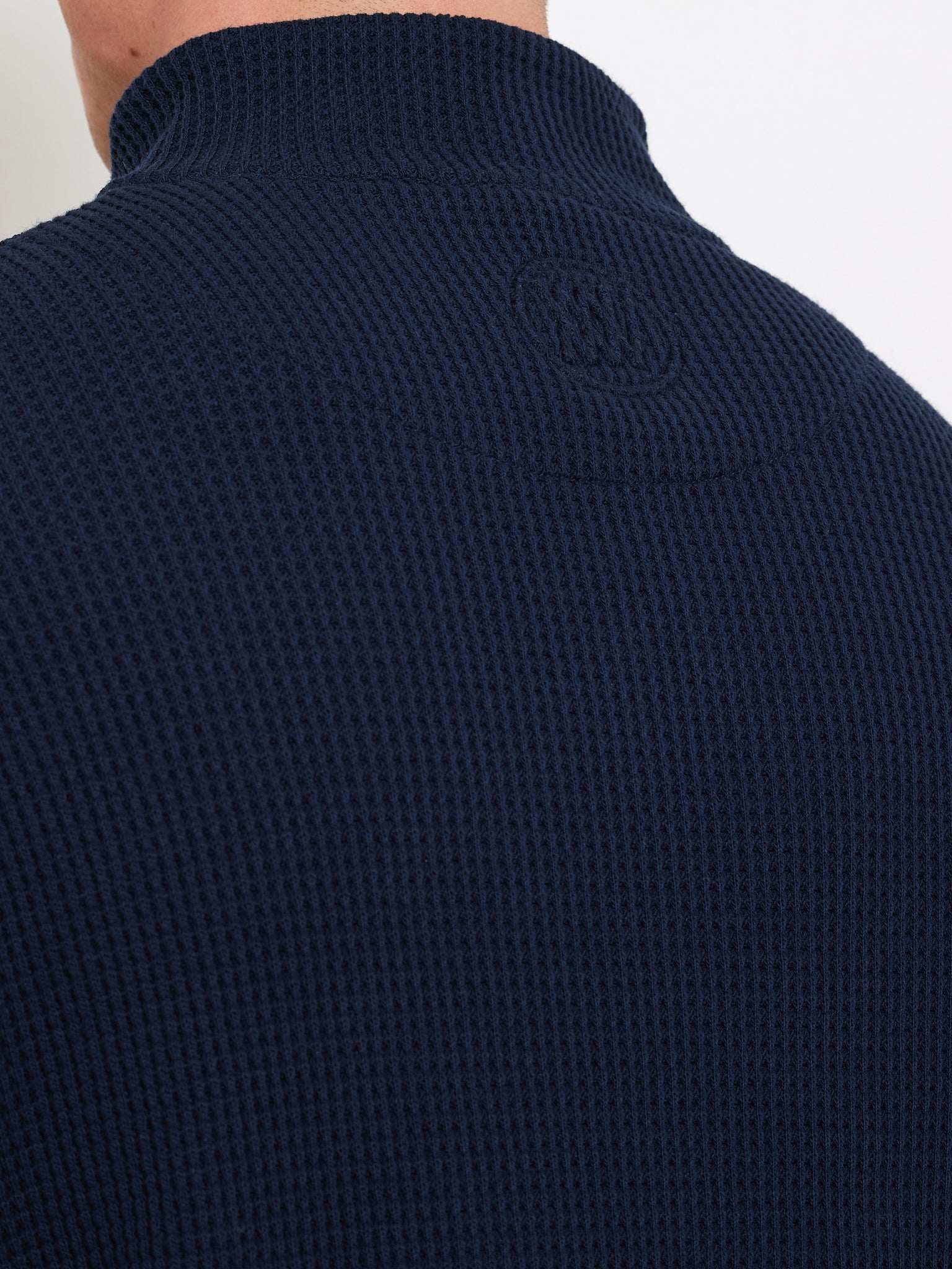Close-up of someone wearing a dark blue Waffle Sweater Men by We Norwegians. This 100% cotton, long-sleeve, high-collar top boasts a waffle-knit pattern and relaxed fit with an embossed logo near the collar. The background is plain white.