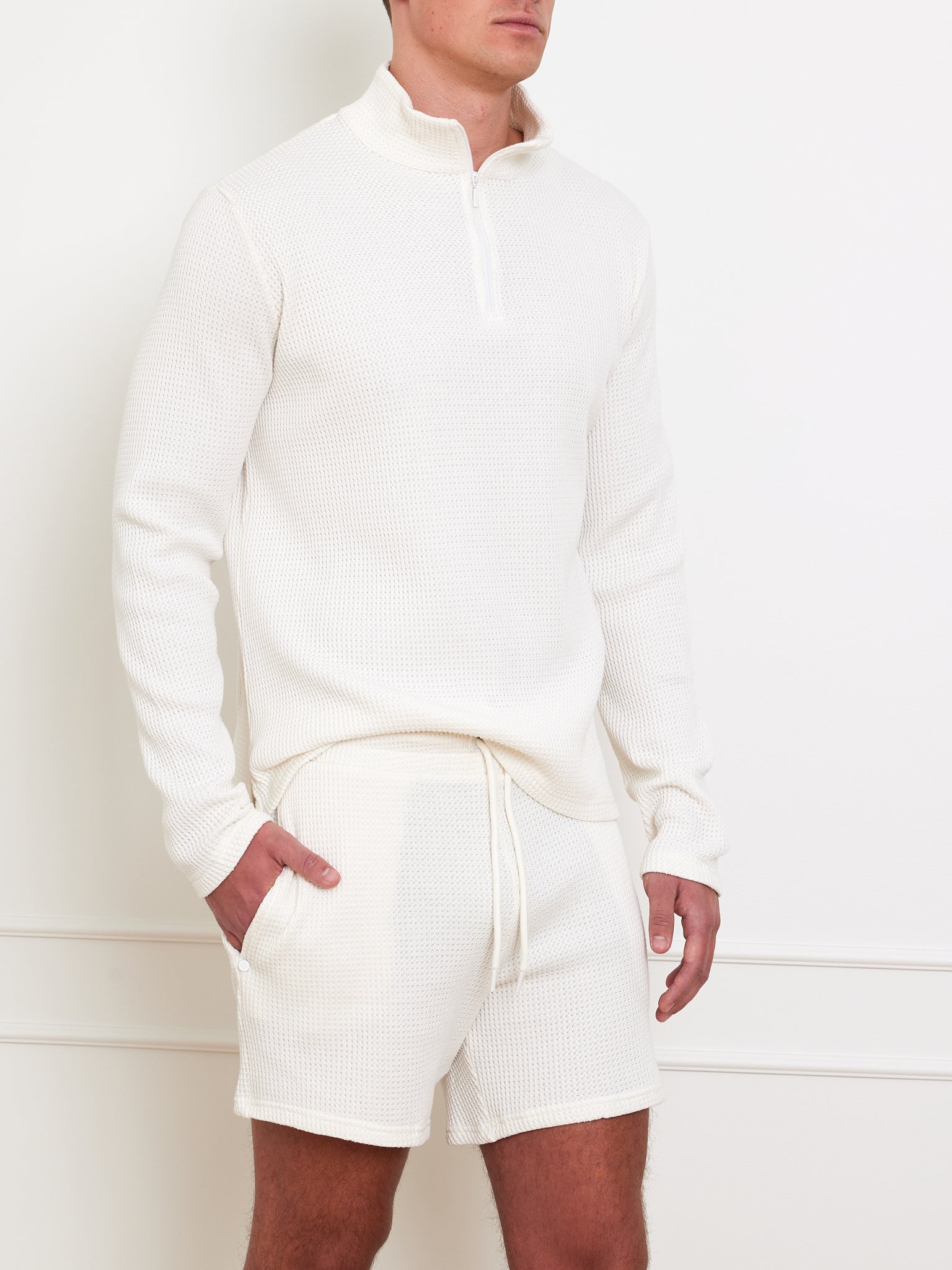 In a neutral room, someone in a We Norwegians Waffle Sweater Men—a white, textured, 100% cotton sweater with a relaxed fit—pairs it with matching shorts. The casual outfit exudes comfort as they stand with hands in the shorts' pockets.