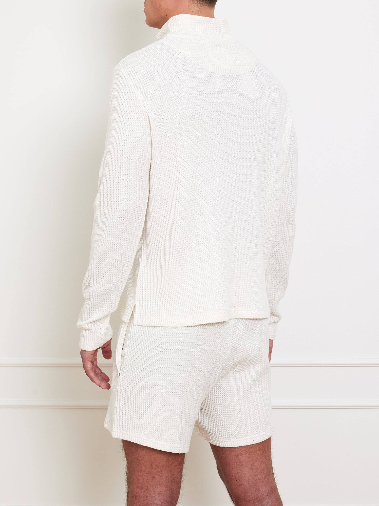 Someone is standing against a plain white wall wearing We Norwegians' Waffle Sweater Men, featuring a relaxed-fit cream-colored waffle-knit long-sleeve top and matching shorts made from 100% cotton for comfort, with their back facing the viewer.
