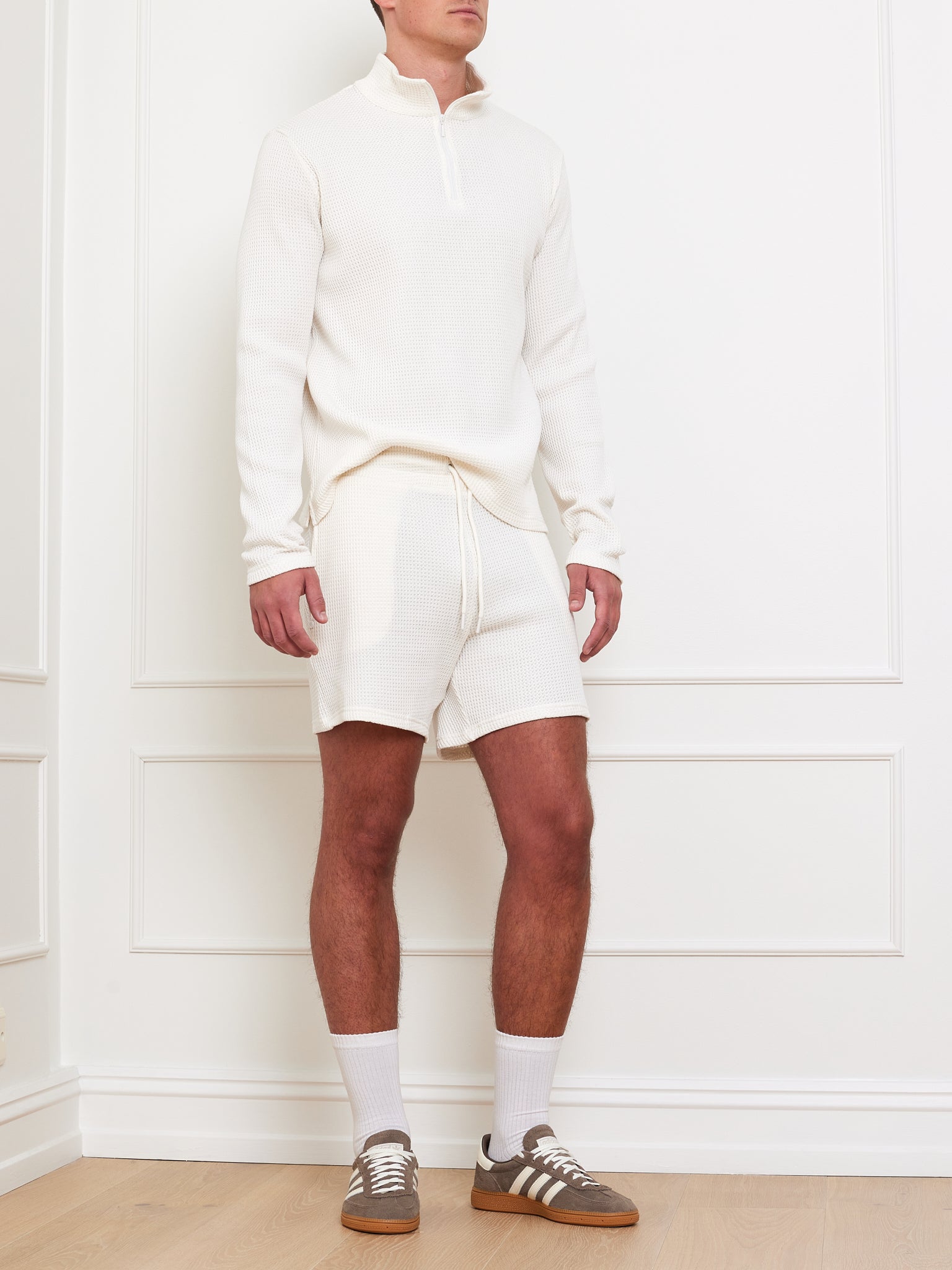In a minimalist room, a person models We Norwegians' relaxed fit featuring the Waffle Sweater Men and 100% cotton shorts. White socks and brown sneakers complete the look against a white wall and light wood floor. The face remains unseen.