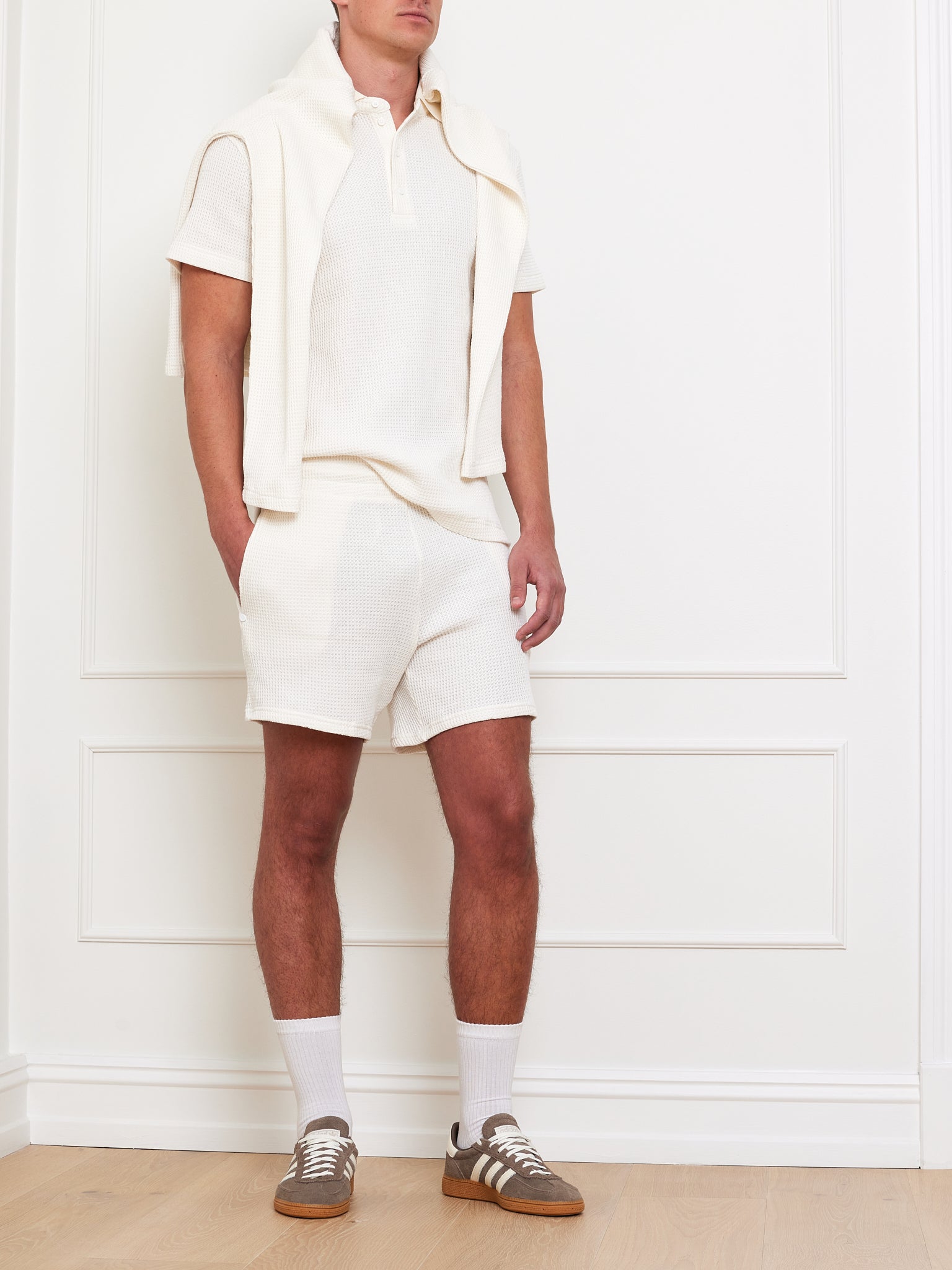 A person stands against a white-paneled wall wearing We Norwegians' Waffle Shorts Men and a white polo, with a sweater draped over their shoulders. They complete the look with white socks, brown sneakers, and one hand in their pocket.