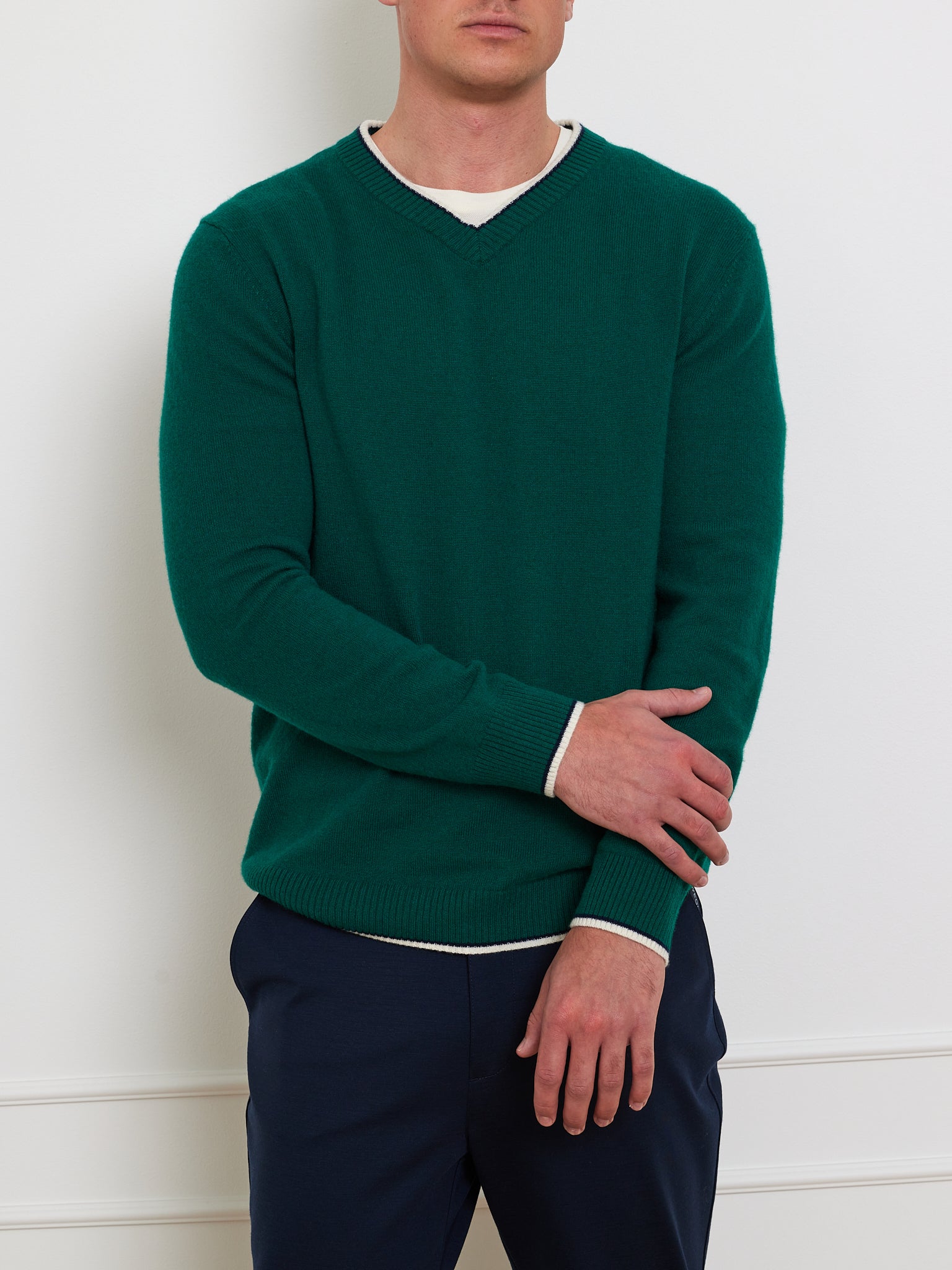 A person leans against a white wall wearing We Norwegians' Club Sweater Men, a green ribbed cashmere V-neck over a white shirt, paired with dark blue pants, and rests one hand on the opposite forearm.