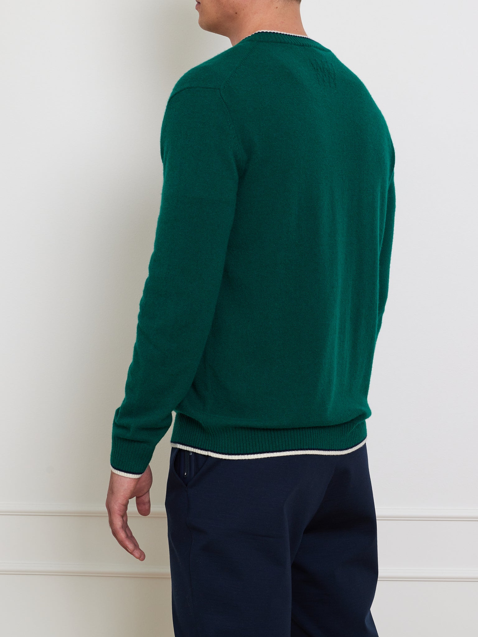 A person facing away from the camera wears We Norwegians' Club Sweater Men, a relaxed-fit dark green cashmere sweater with a ribbed V-neck, paired with navy blue pants. Their hand rests casually against a plain, light-colored wall.