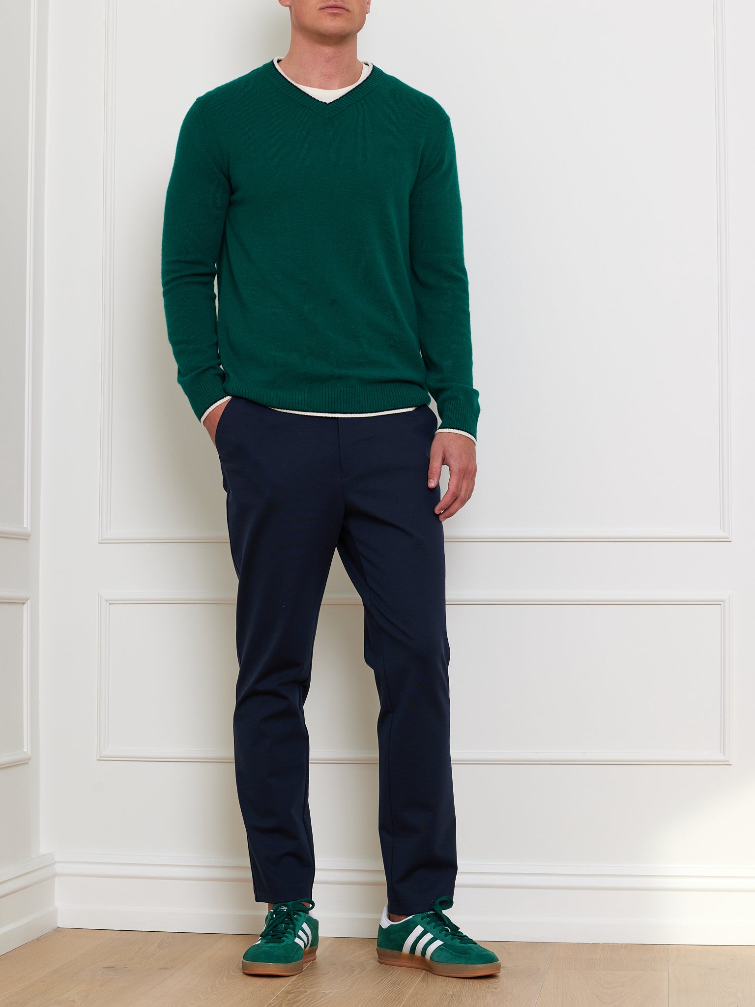 A person in a We Norwegians Club Sweater Men, a relaxed fit green cashmere with a ribbed V-neck, and navy trousers stands against a white paneled wall. They wear green and white sneakers, one hand in their pocket, on light wood flooring.