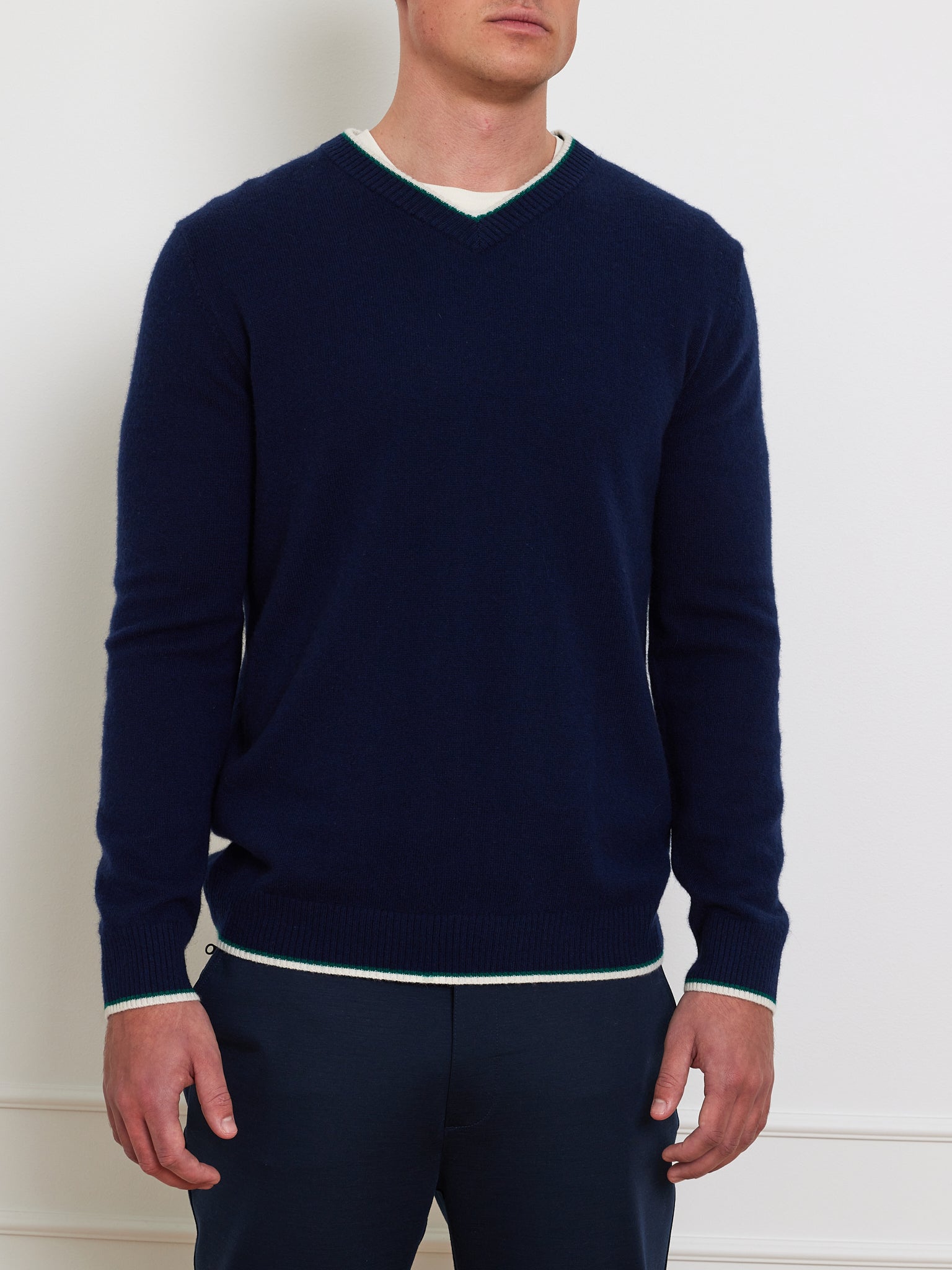 Wearing the Club Sweater Men by We Norwegians, a person stands against a plain white wall. The relaxed-fit navy blue ribbed V-neck is layered over a white shirt and paired with dark trousers, their face remains unseen.