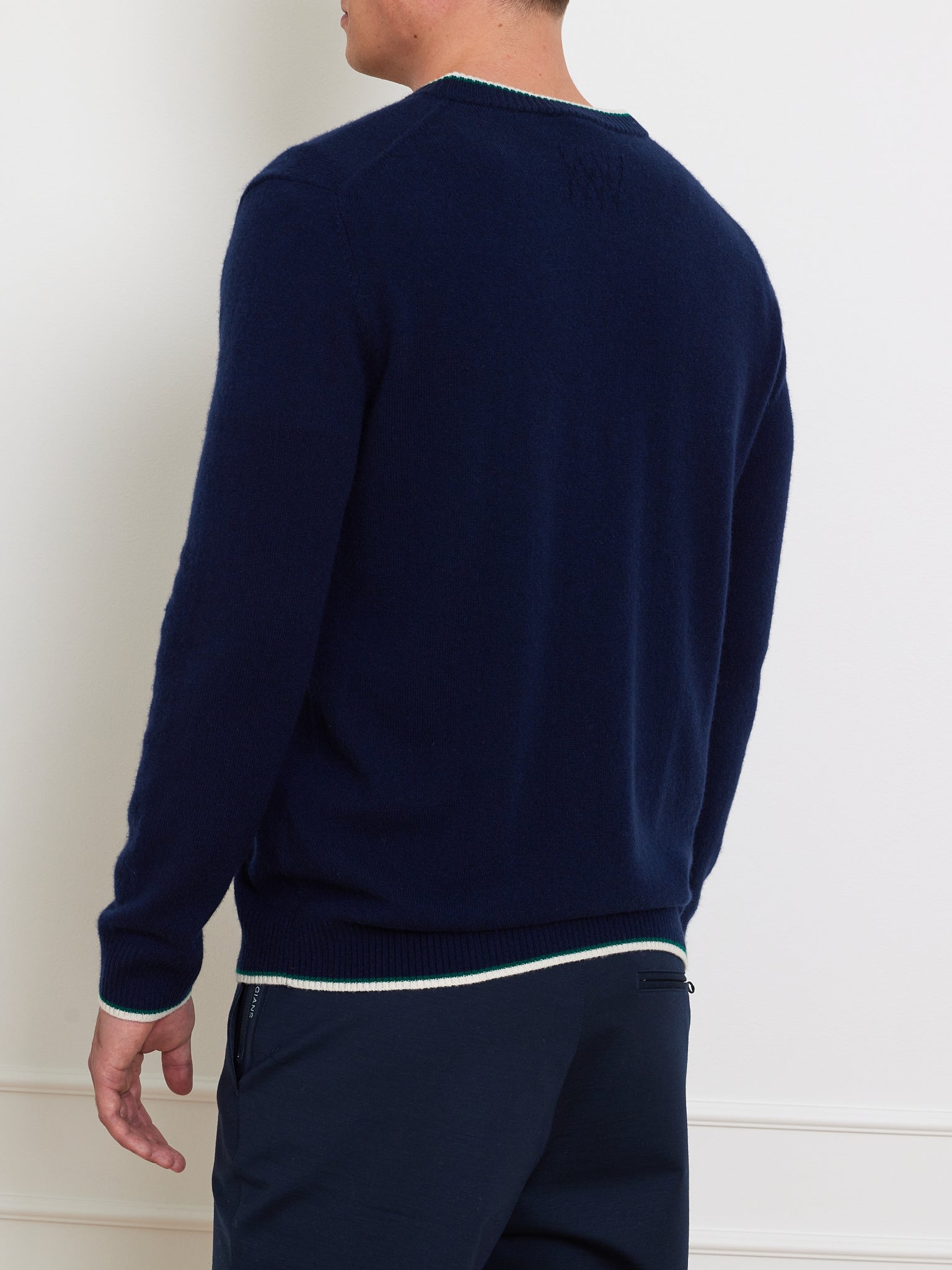 A person is wearing the We Norwegians Club Sweater Men, a relaxed fit in dark blue cashmere with light trim, along with dark pants, standing facing away from a white wall.