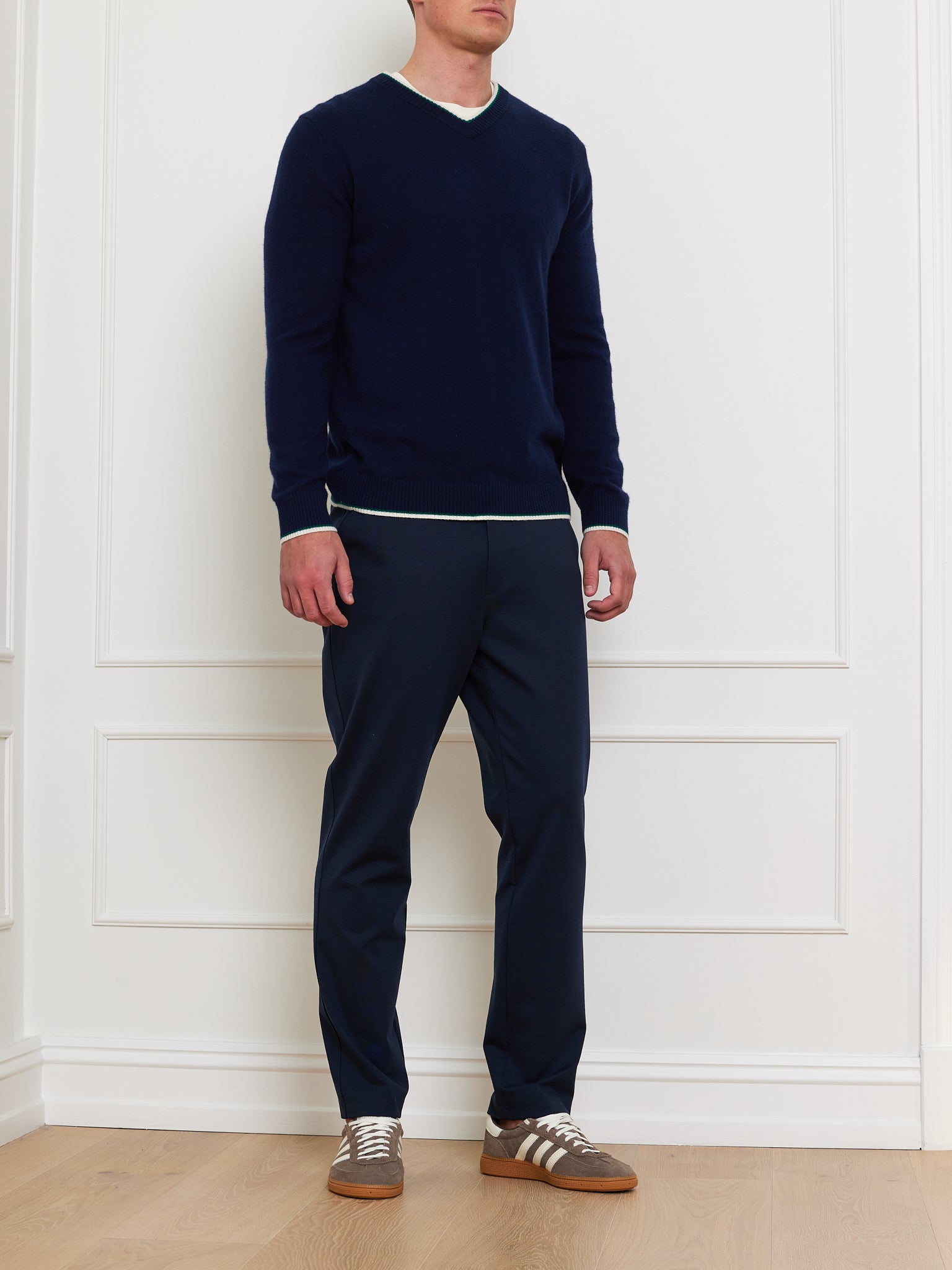 A person wears a We Norwegians Club Sweater Men, featuring a relaxed fit in dark blue with a ribbed V-neck, paired with navy pants and brown sneakers, while standing against a white wall on a wooden floor.