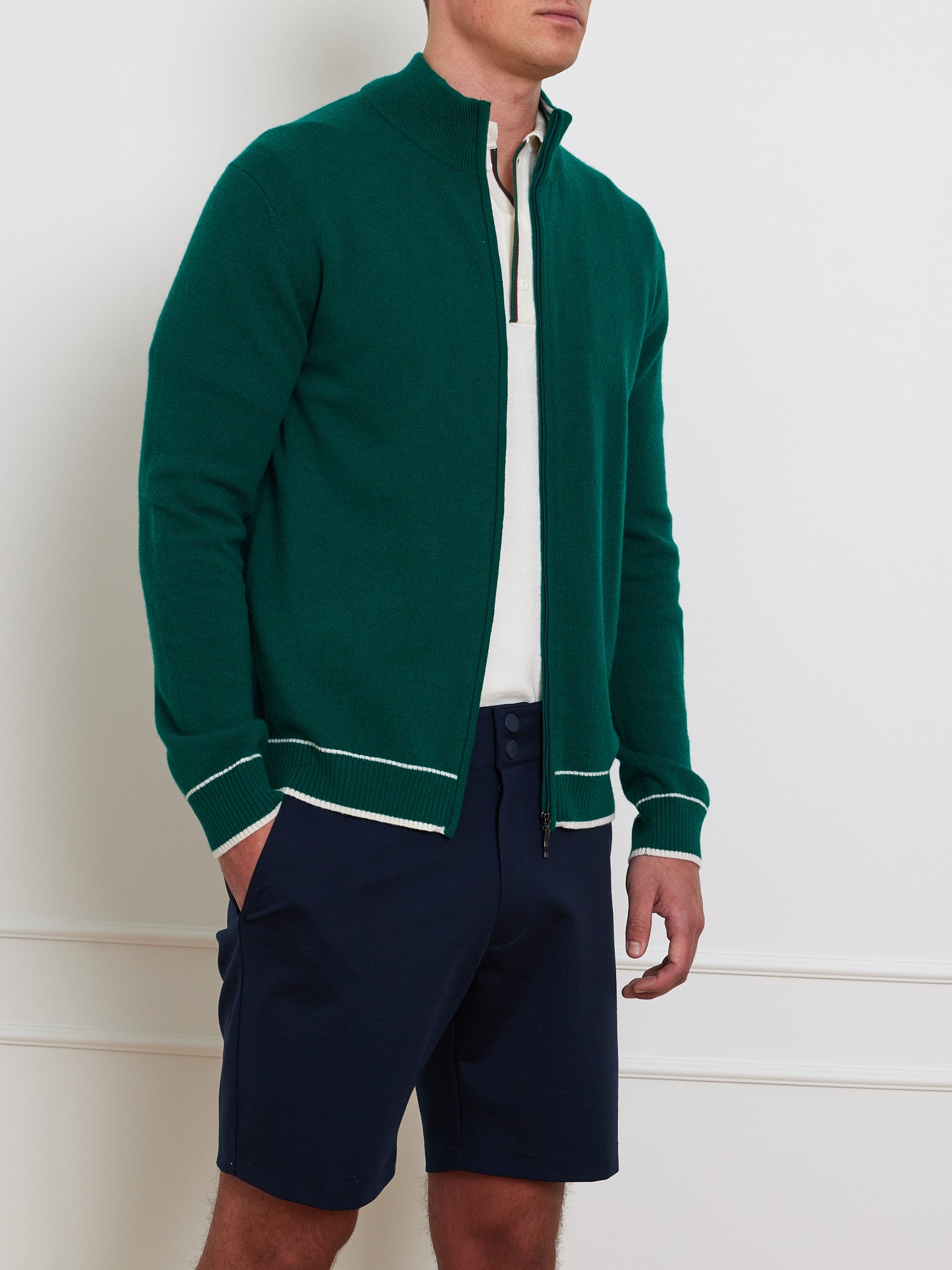 A man embodies an après sport style wearing the Club Cardigan Men by We Norwegians, a green zip-up sweater that evokes luxury sportswear, over a white shirt and navy blue shorts. He stands with one hand in his pocket against a plain white background.