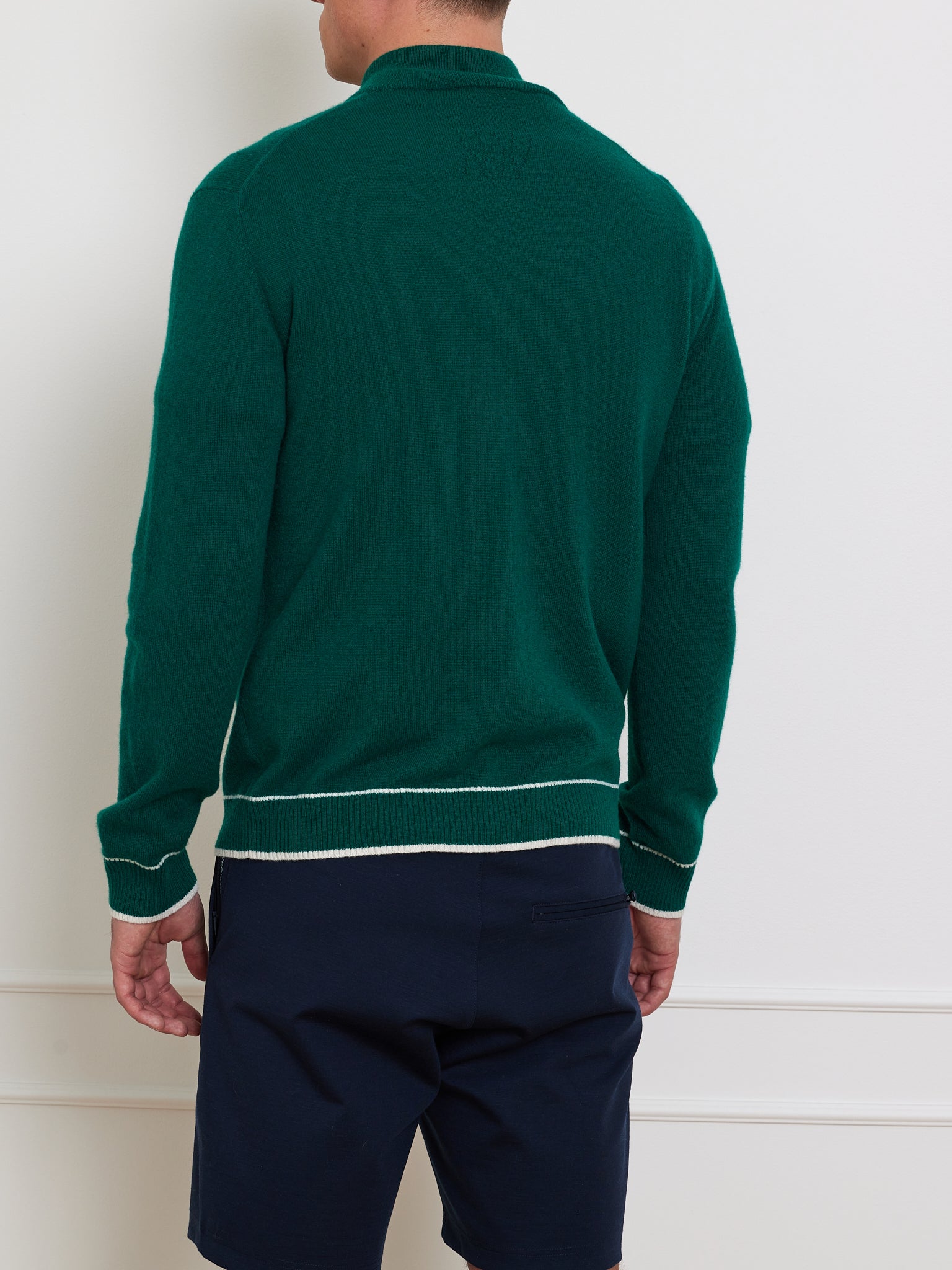 A man in a Club Cardigan Men by We Norwegians, featuring green with white trim, pairs it stylishly with dark shorts as he stands confidently against a plain white wall, showcasing a blend of casual elegance and après sport flair.