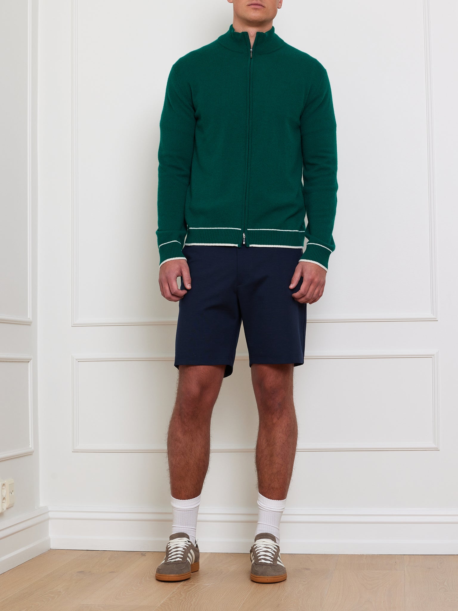 In a white paneled room, a person showcases an après sport vibe with We Norwegians' green Club Cardigan Men zip-up, navy shorts, white socks, and gray sneakers. The wooden floor offers a warm contrast while the face stays unseen, highlighting the casually refined ensemble.