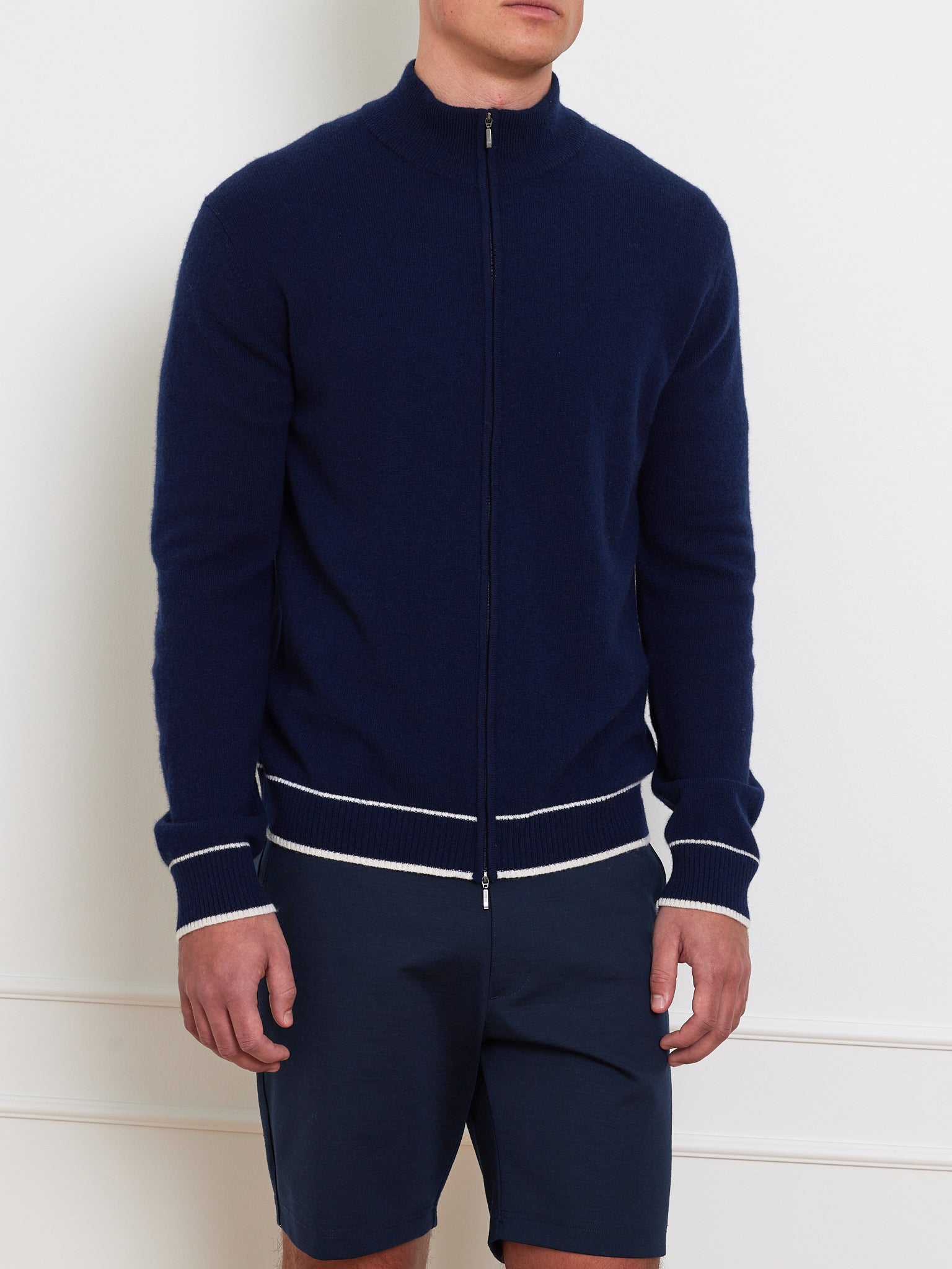 Someone is photographed in an elegant après sport style, wearing We Norwegians' Club Cardigan Men in luxurious navy cashmere with white trim, paired with matching navy shorts, against a plain white wall that conceals their identity.