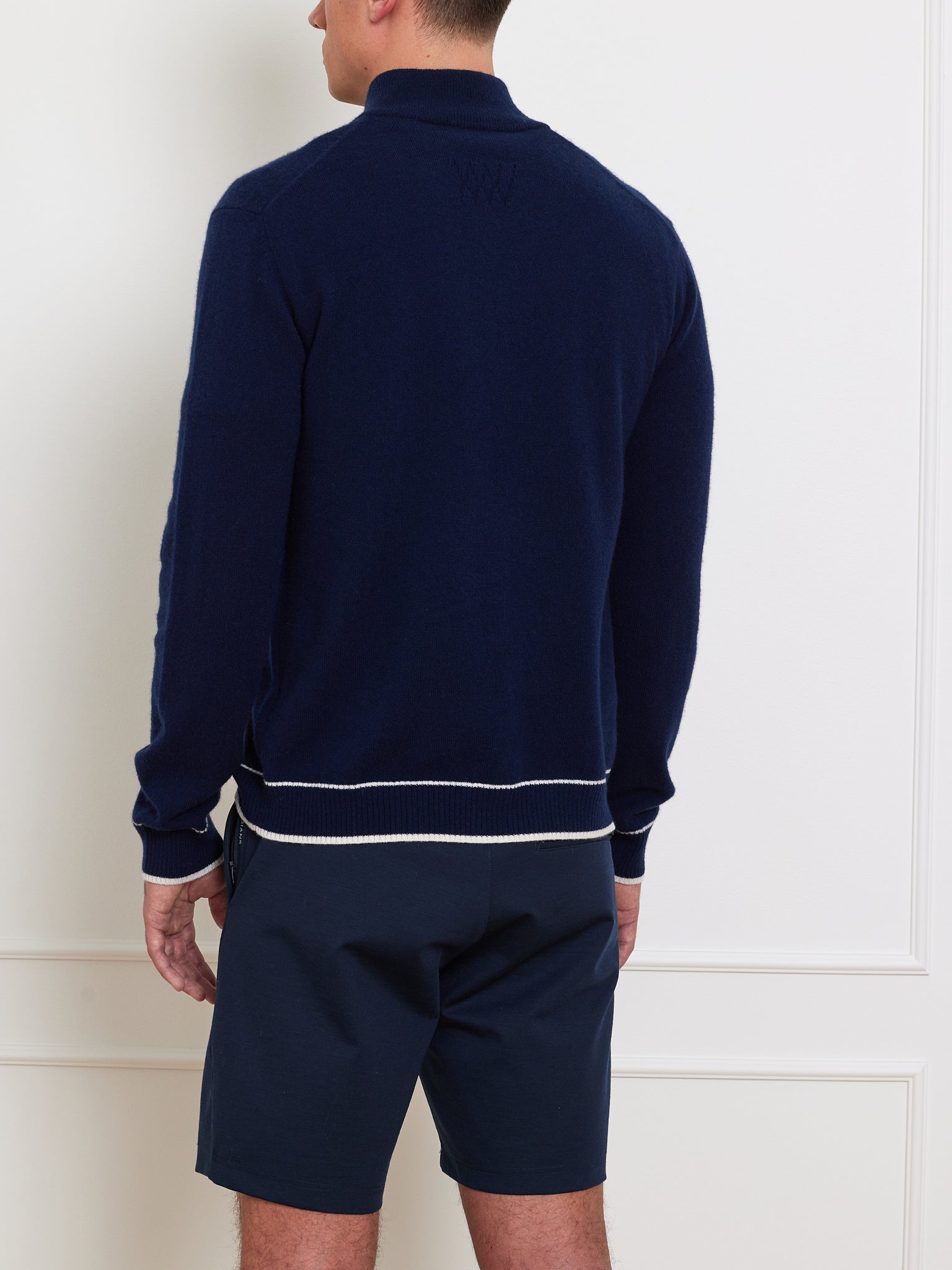 A person models an après sport look in front of a plain white wall, wearing We Norwegians' Club Cardigan Men, a dark blue long-sleeve sweater with white trim, paired elegantly with navy shorts, embodying the essence of luxury sportswear.