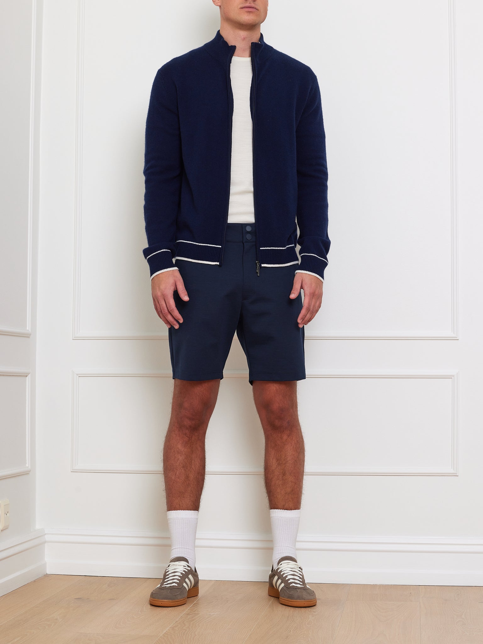 A man in a navy We Norwegians Club Cardigan, white shirt, navy shorts, white socks, and brown sneakers exudes an après sport look against a white paneled wall on a wooden floor.