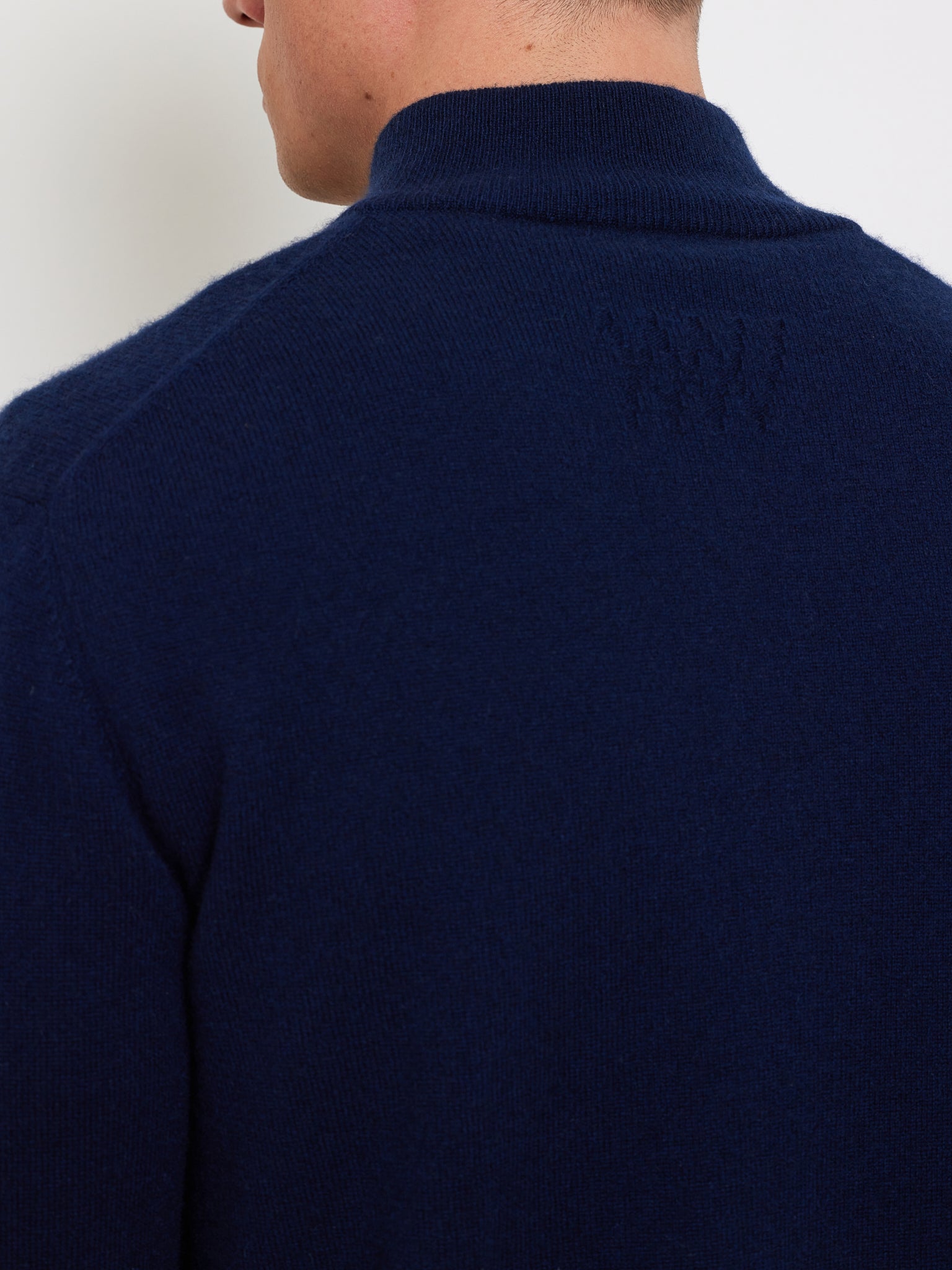 A person facing away, wearing the "Club Cardigan Men" by We Norwegians, in dark blue with a high collar and textured upper back details. This garment combines luxury sportswear with elegance for a refined après-sport look focused on the back and shoulders.