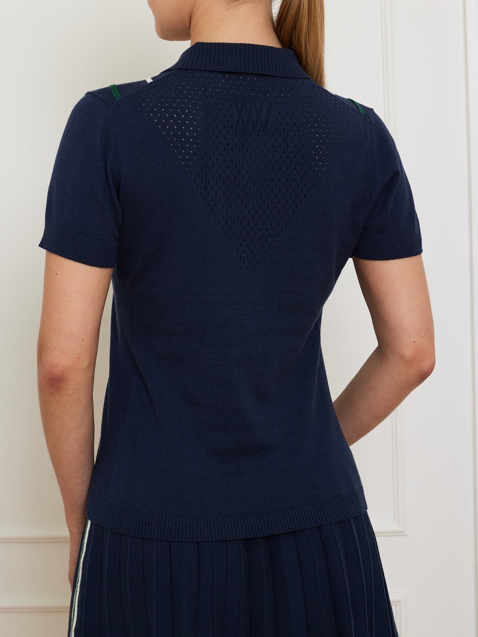 A person in a navy Team Chevron Polo by We Norwegians, featuring a breathable mesh triangle back design, stands against a white wall. The top is paired with a pleated skirt for an ideal style-comfort blend.