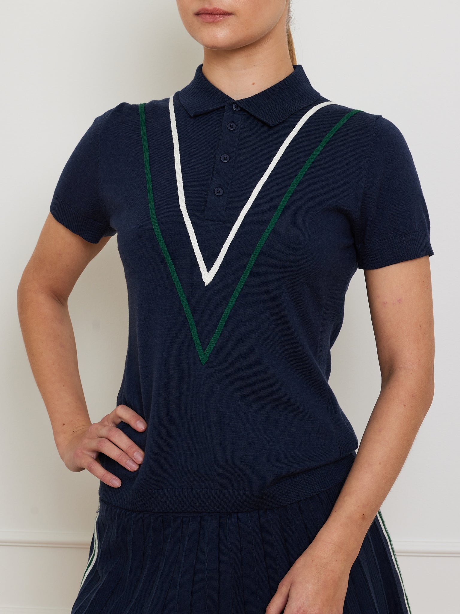 A person wears the Team Chevron Polo Women by We Norwegians, made of breathable mesh. Their left hand is on their hip against a plain light wall. The navy polo pairs stylishly with a pleated skirt, combining comfort and modern flair.