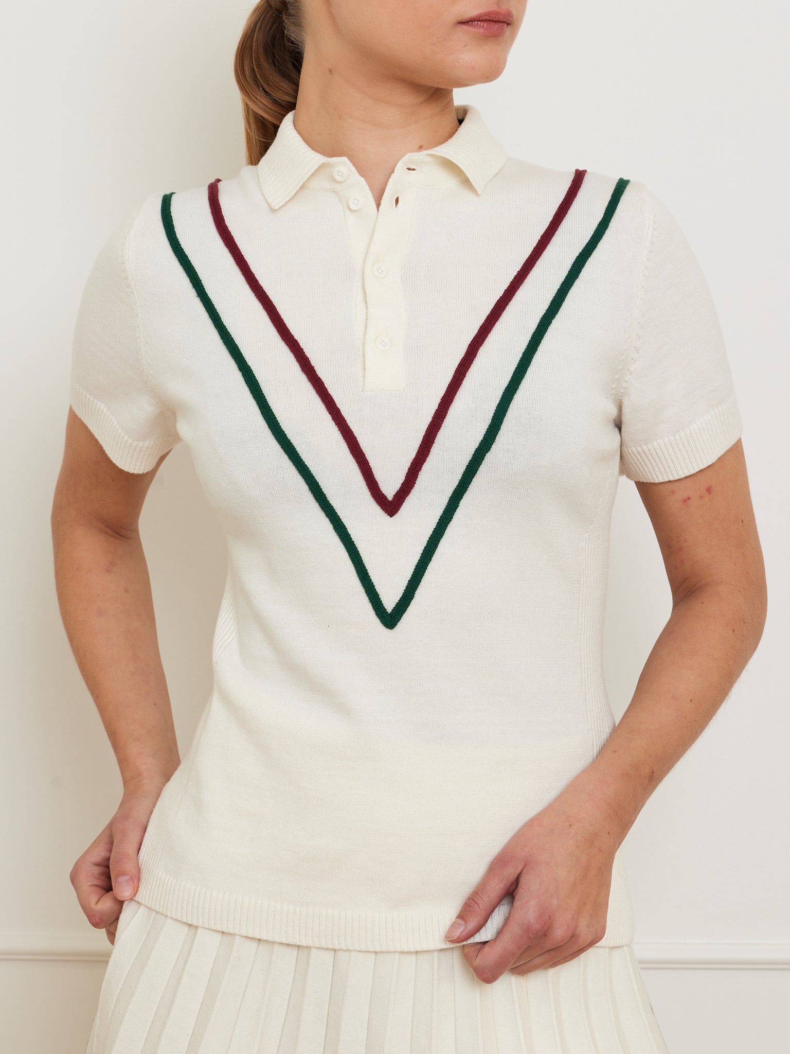 Wearing the We Norwegians Team Chevron Polo Women, a person strikingly poses with one hand on their hip, dressed in a white, short-sleeved knit featuring red and green chevron stripes and a collar. The outfit is paired with a pleated skirt against a simple, light backdrop.