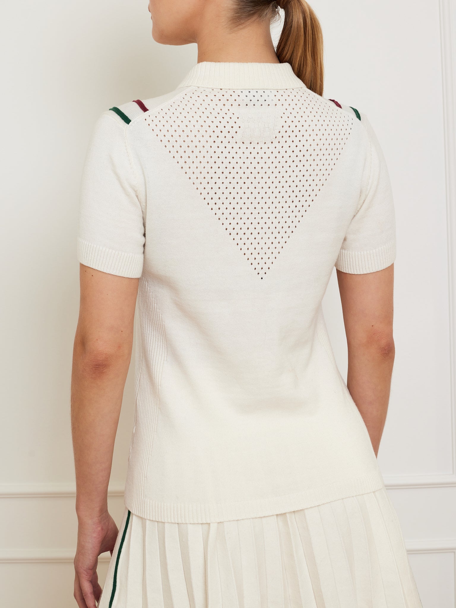 A person with a ponytail stands against a light-colored wall wearing the Team Chevron Polo Women by We Norwegians. This white merino cotton top features short sleeves, perforated triangles on the back, and green and red shoulder accents, offering breathable mesh comfort.