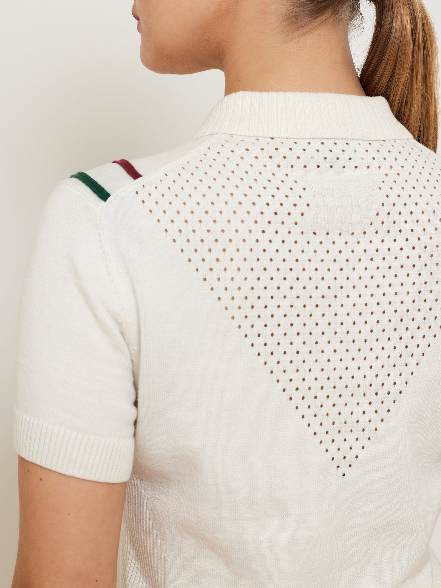 A person, turned slightly away from the camera, is wearing We Norwegians' Team Chevron Polo Women—a white, short-sleeved merino cotton knit sweater with a perforated triangle pattern on the back, an embroidered logo, and shoulder stripes.