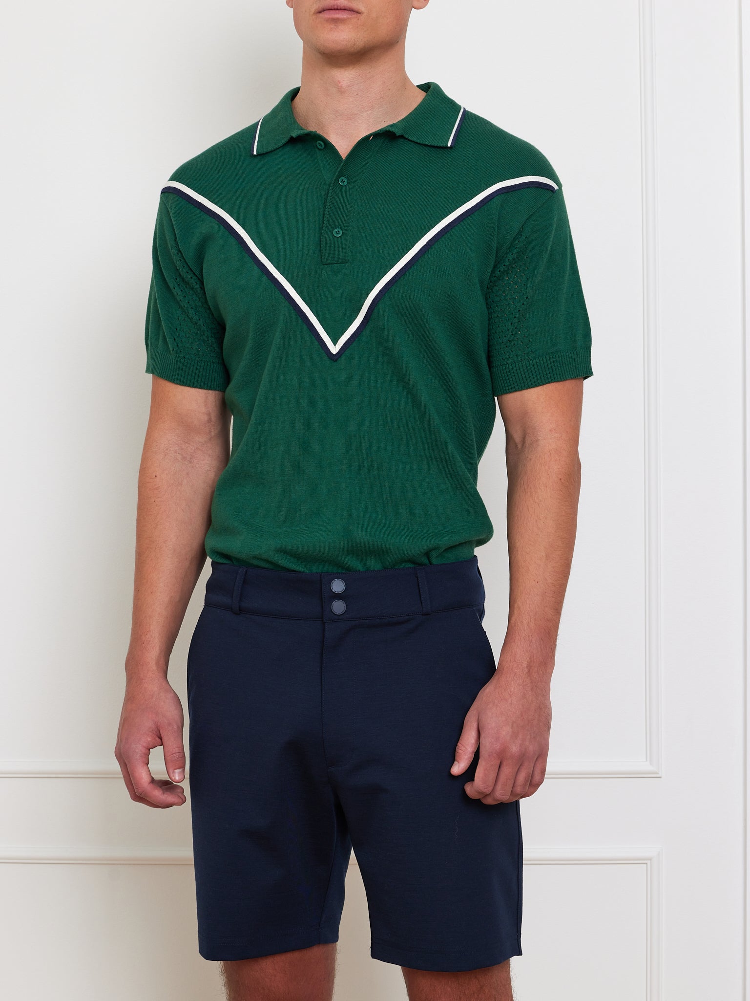 Wearing We Norwegians' Team Chevron Polo Men in a merino-cotton blend, someone stands against a white wall. The polo's distinctive white chevron design pairs effortlessly with navy blue shorts, featuring a collar and button placket that emphasize performance-driven style, while their head is unseen.