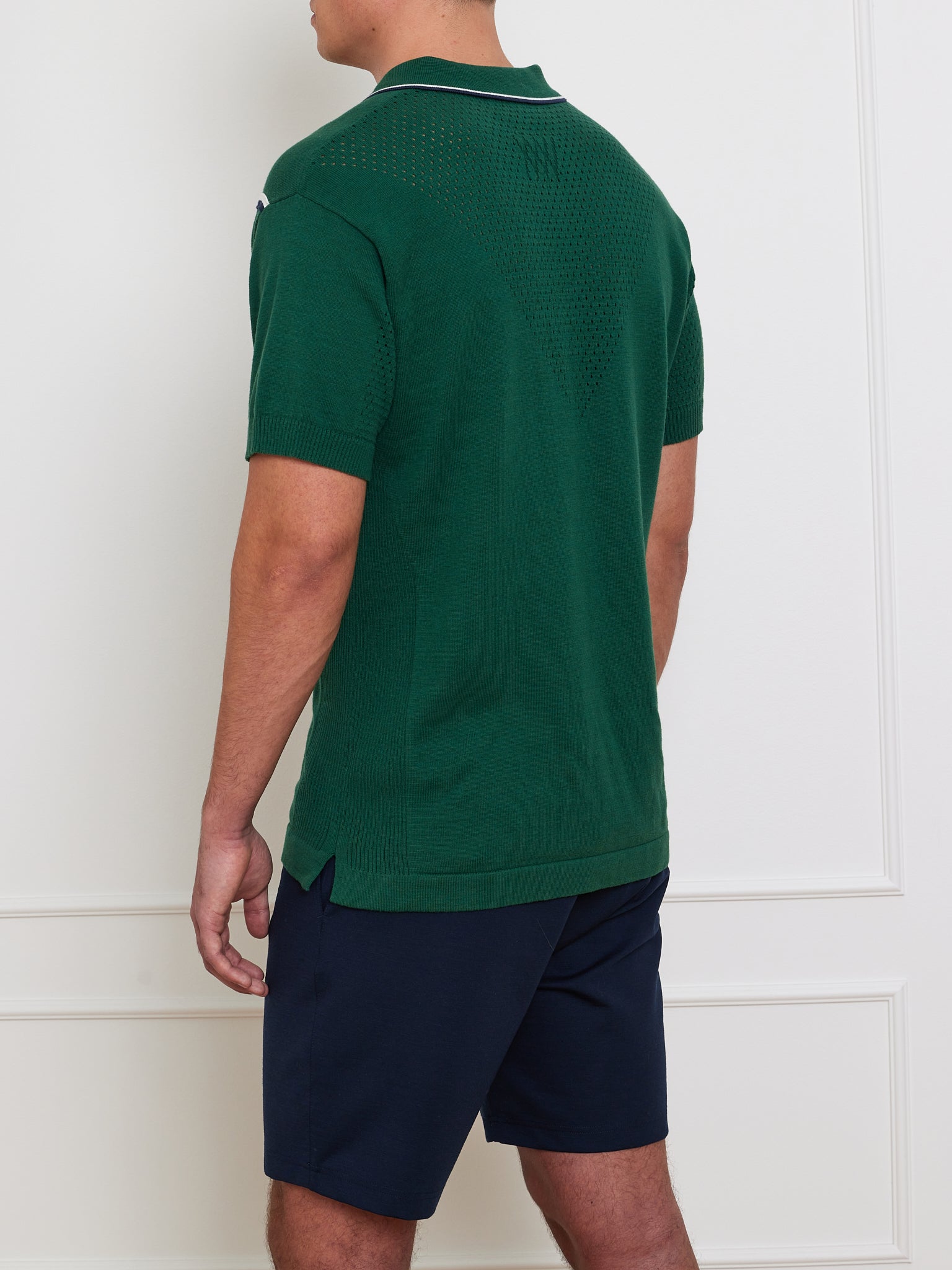 A person stands against a white wall, wearing the We Norwegians Team Chevron Polo in green and dark navy shorts. The focus is on the polo's back, showcasing texture details and a small collar logo, crafted from a merino-cotton blend for performance-driven style.