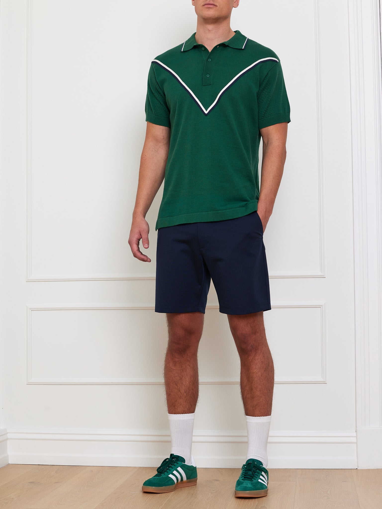 A person wearing a We Norwegians Team Chevron Polo with a white V-pattern, navy blue shorts, white socks, and green Adidas sneakers with white stripes stands in a room with white paneled walls and a wooden floor.