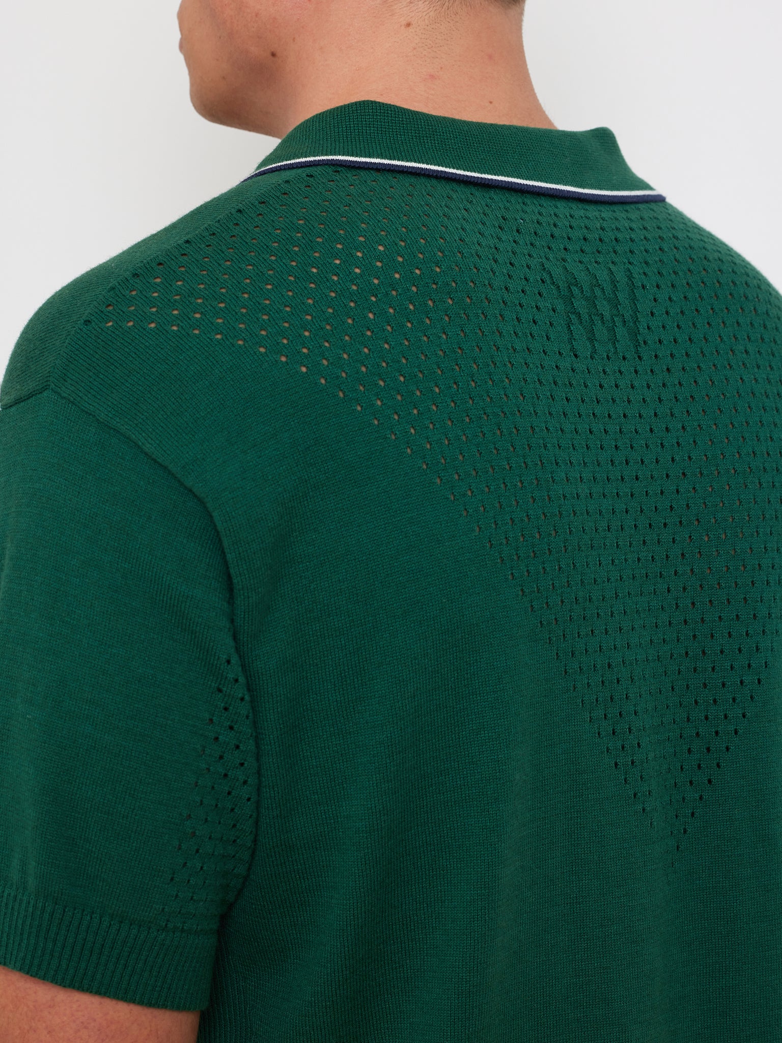 A person is seen from the back wearing a green We Norwegians Team Chevron Polo Men, featuring short sleeves, a performance-driven perforated knit with a textured pattern, and a thin white stripe on the collar for added flair.