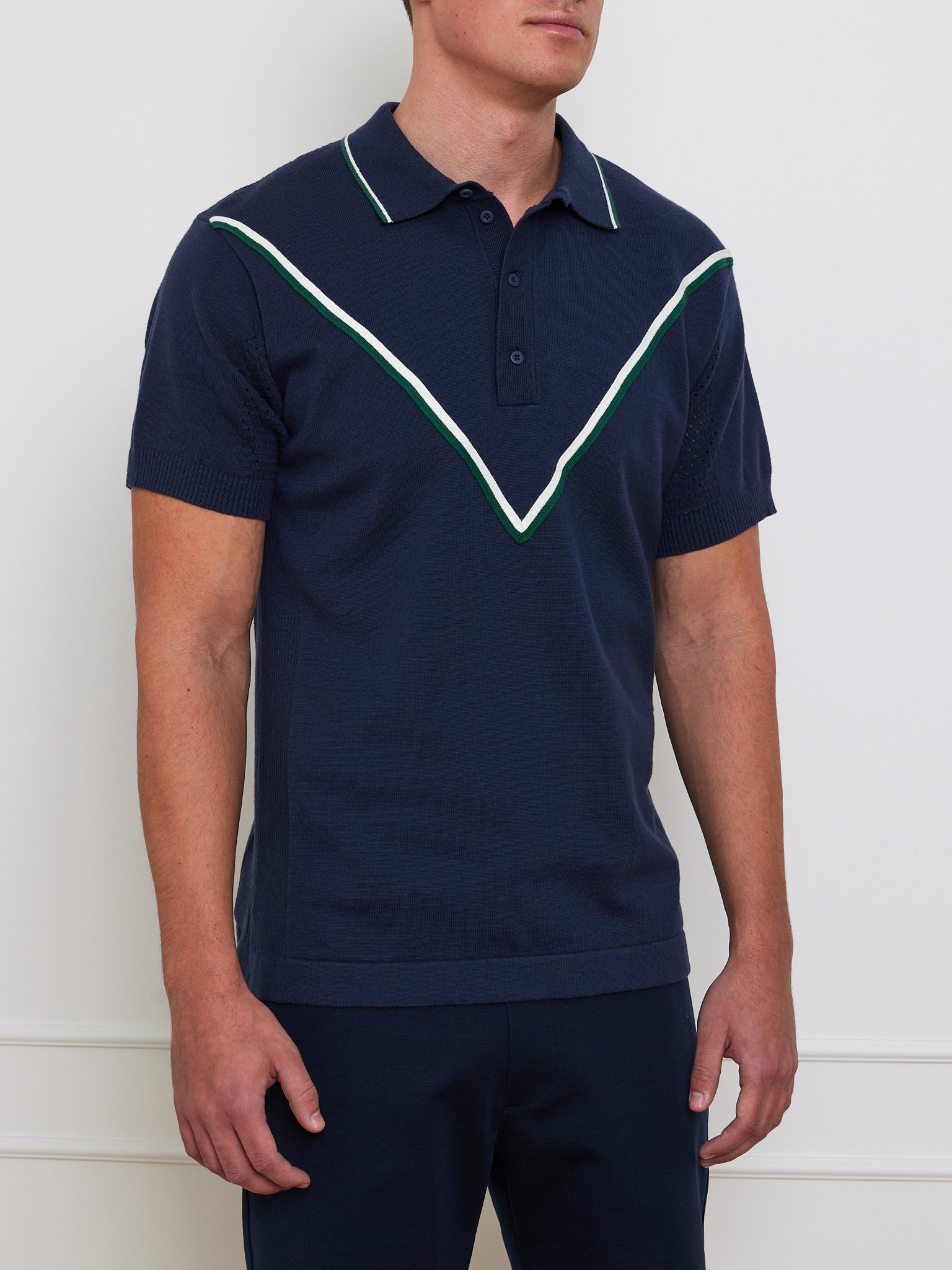 A person wears the We Norwegians Team Chevron Polo Men, a navy merino-cotton blend with a performance-driven style featuring a white and green chevron chest design. The shirt has a classic collar and short sleeves, showcased against a white wall.