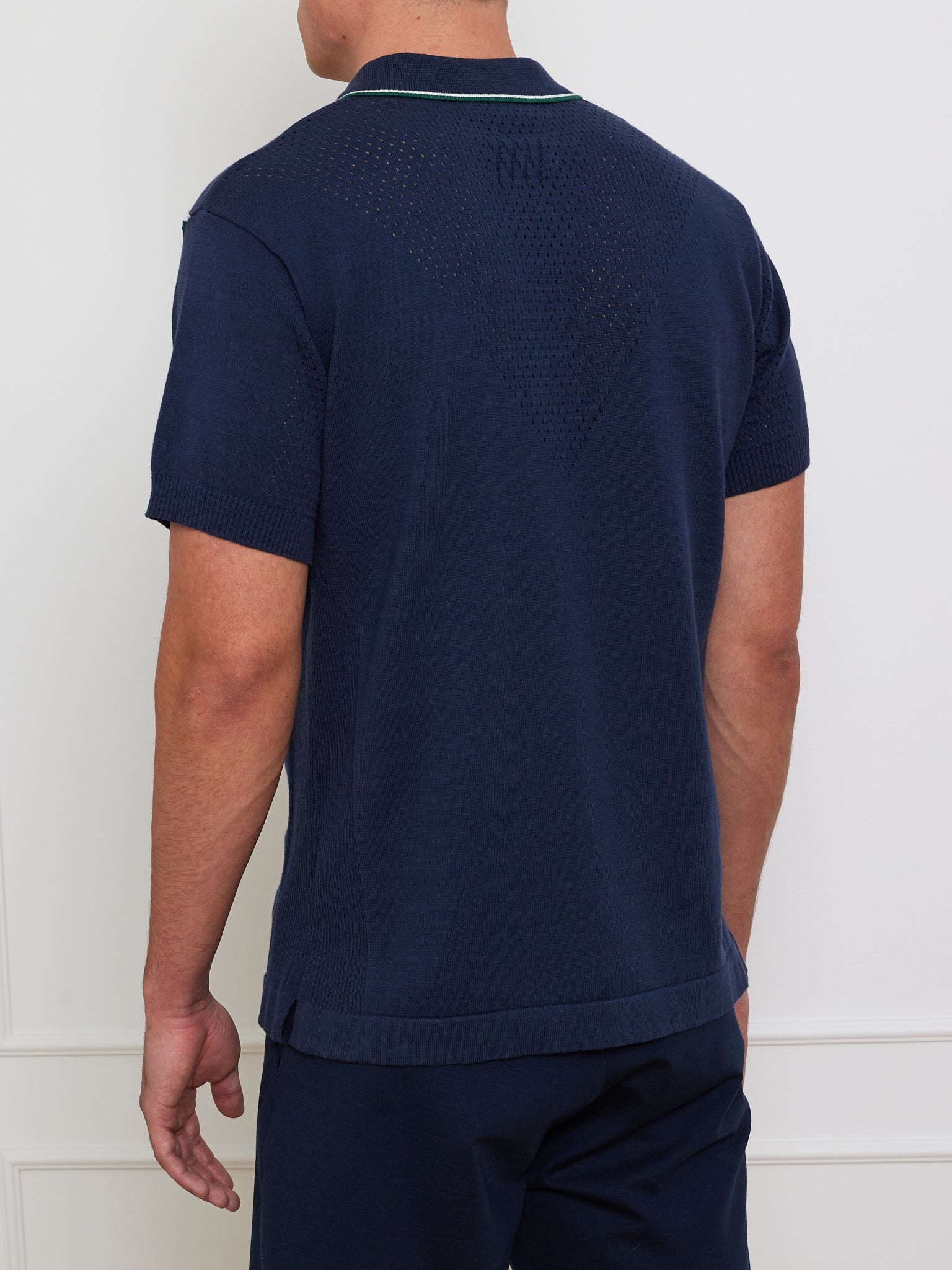 A person wearing a Team Chevron Polo from We Norwegians is seen from behind. The navy blue knit shirt has a textured back and short sleeves, paired with matching pants. They stand indoors against a white wall backdrop.