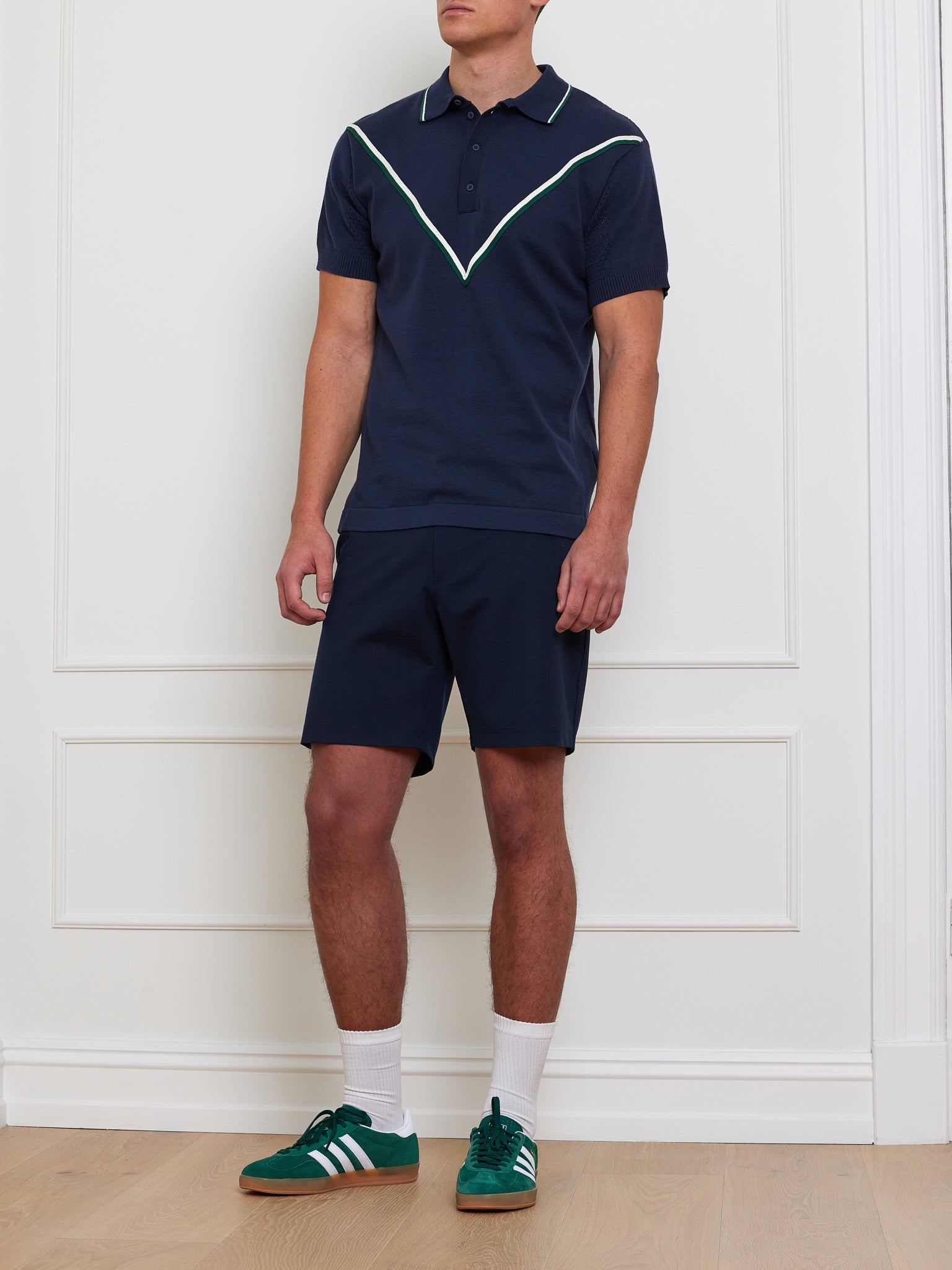 Indoors, a person dons a We Norwegians Team Chevron Polo in navy with white piping, paired with matching shorts. They wear green sneakers with white stripes and socks. The backdrop is a panel-detailed white wall, enhancing the performance-driven vibe. Their face isn't shown.