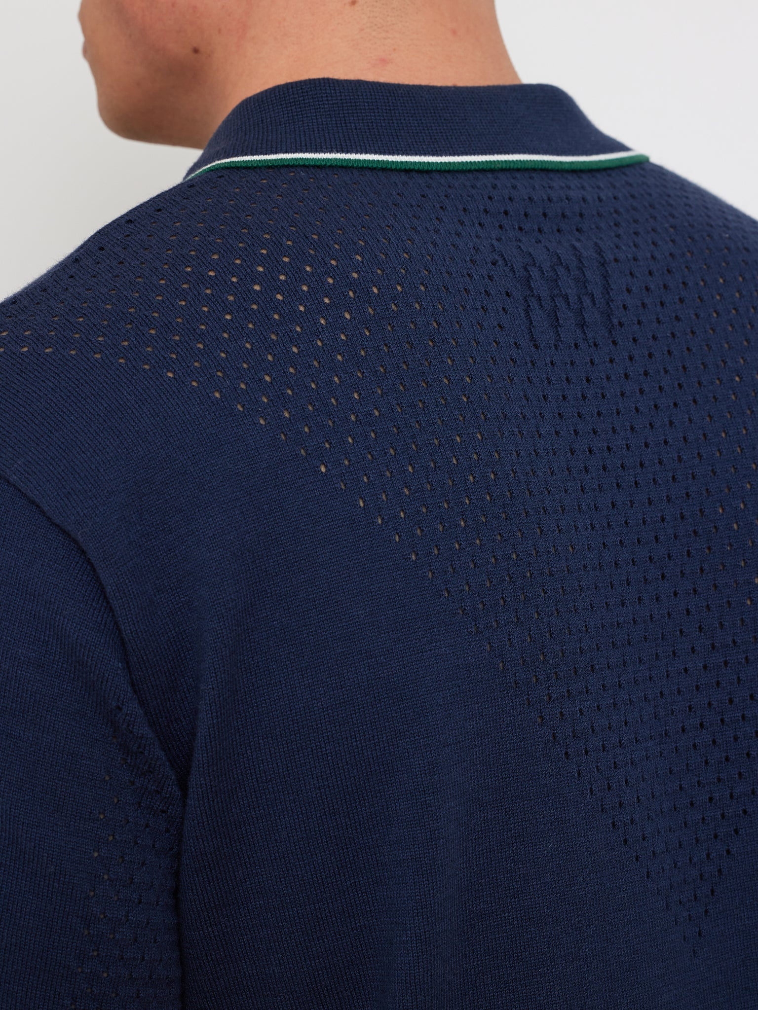 Close-up of a person in the We Norwegians Team Chevron Polo Men, featuring a navy blue performance-driven style with perforated pattern, and a neckline accented by thin white and green stripes. The merino-cotton blend is highlighted from the side and back view, with neck and hairline visible.