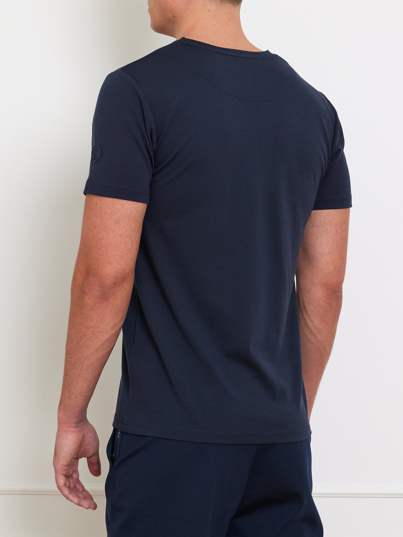 A person is seen from behind in a fitted navy blue Medalist Piqué Tee Men by We Norwegians, made of CoolMax blend, paired with matching pants. The short-sleeved tee features a small round emblem on one sleeve and they stand against a light-colored wall.