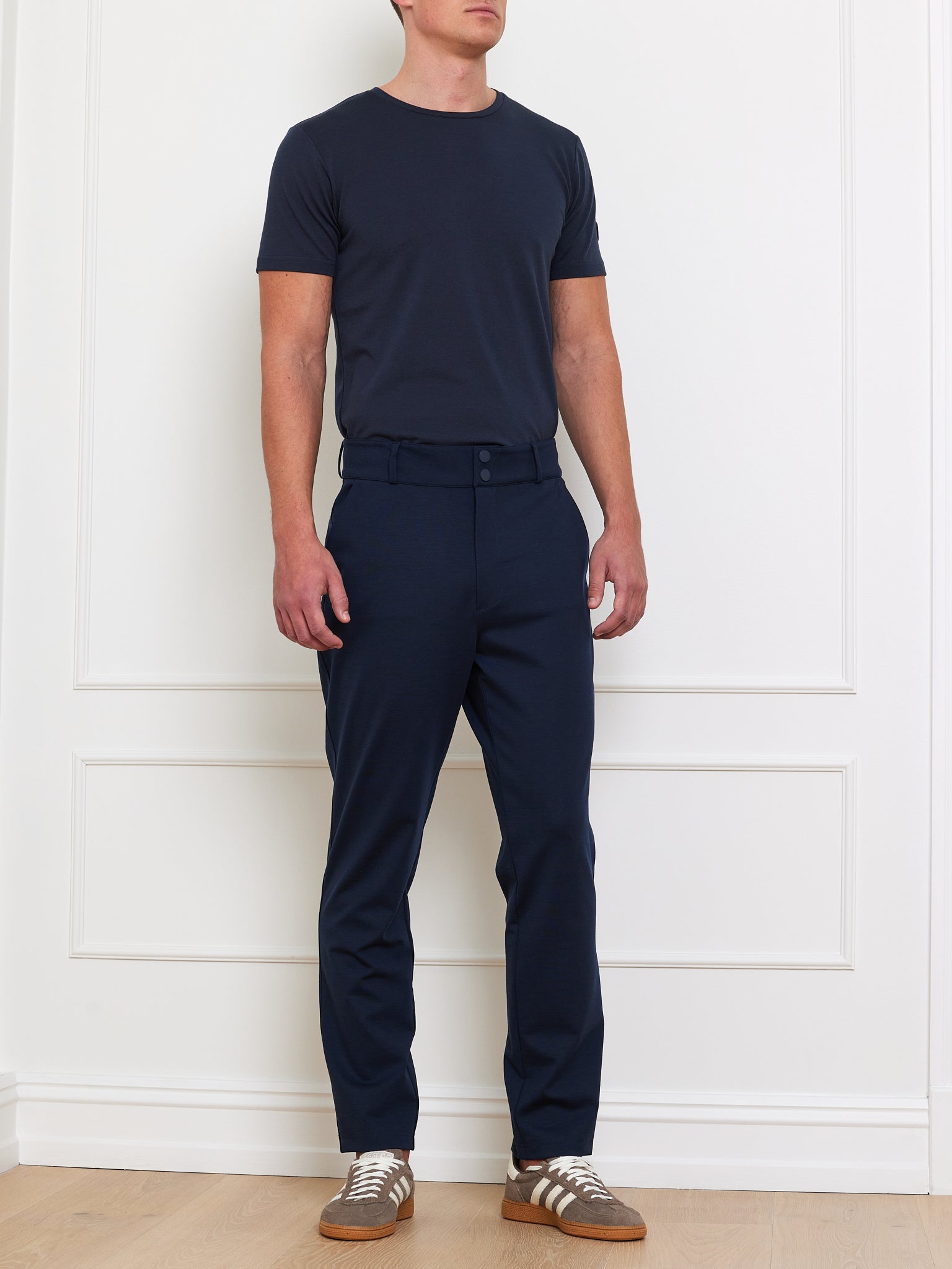 A person is against a white wall, wearing a We Norwegians Medalist Piqué Tee Men with matching pants and belt. Brown sneakers with white laces complete the look. The setting features a wooden floor and white paneling for contrast.