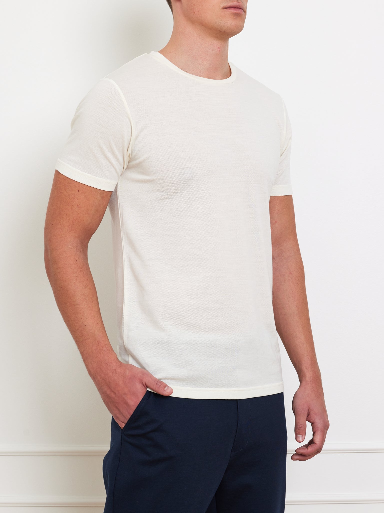 A man stands against a light wall wearing the We Norwegians' Medalist Piqué Tee Men and dark pants, one hand in his pocket.