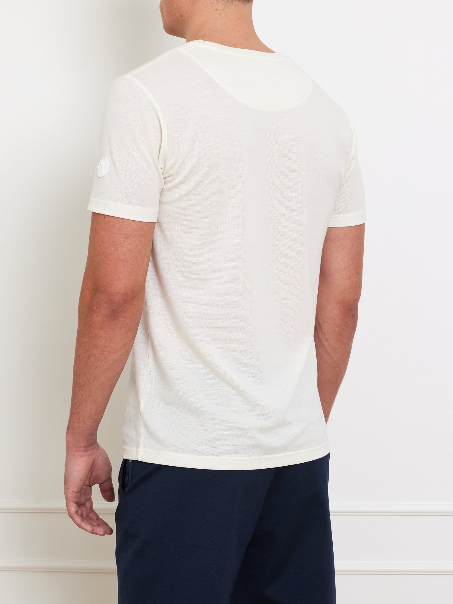 A person in a We Norwegians Medalist Piqué Tee Men and navy blue pants stands against a white wall, facing away from the camera.