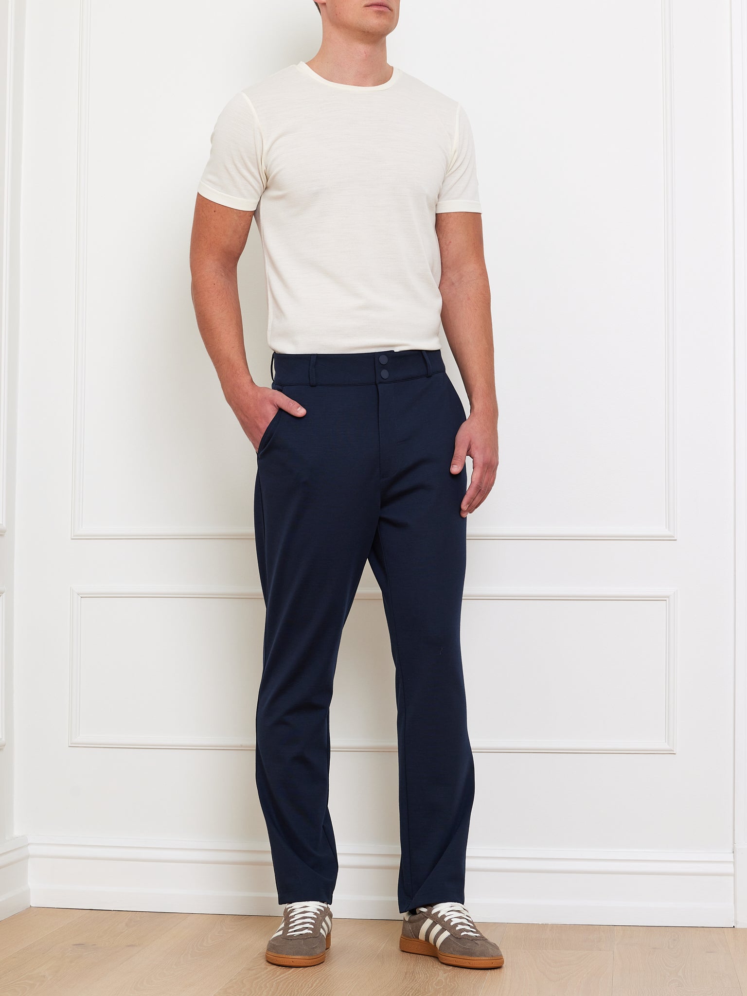 A man in a We Norwegians Medalist Piqué Tee men’s T-shirt, dark blue pants, and brown sneakers with white laces stands casually with his hands in his pockets against a white wall.