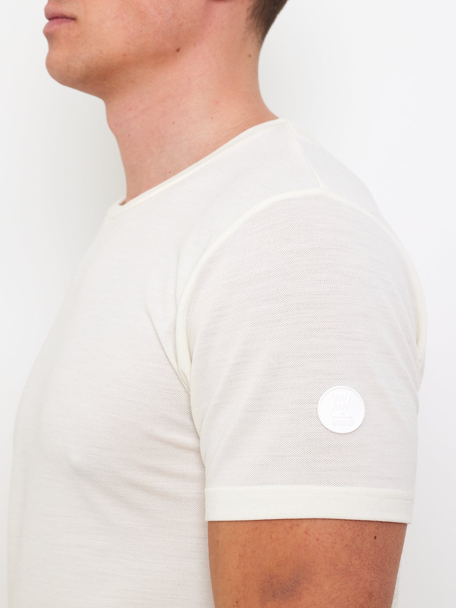 Side profile of a person wearing the Medalist Piqué Tee Men by We Norwegians, a light-colored, short-sleeved golf T-shirt made from piqué merino wool with a small circular logo on the sleeve. The garment's sleek design and premium fabric are highlighted against a white backdrop.