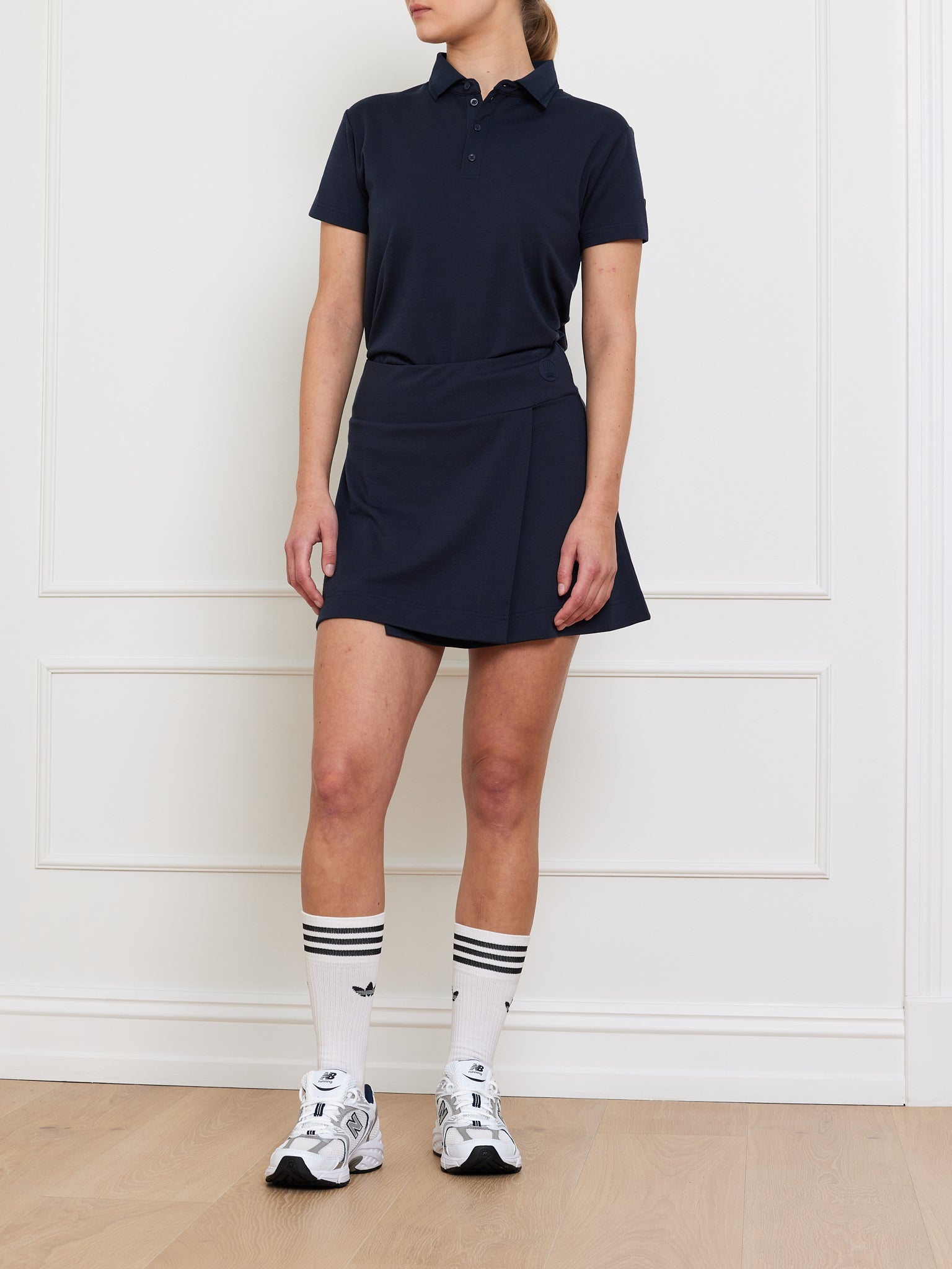 Dressed in a navy polo and the Medalist Piqué Skirt Women by We Norwegians, a person stands against a white wall. Their outfit is completed with white sneakers and knee-high socks featuring black stripes.