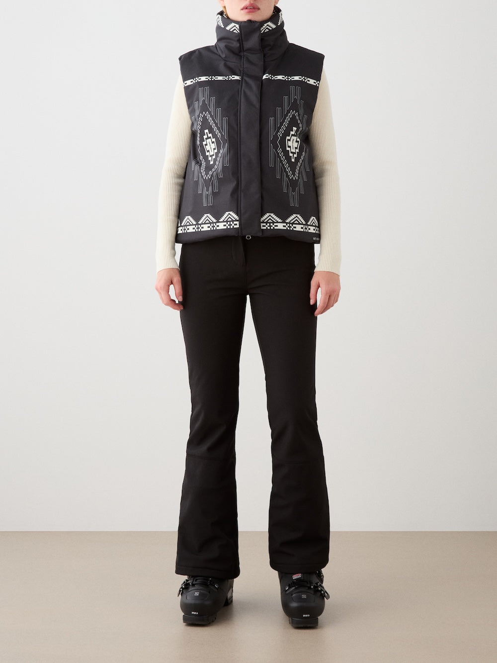 A woman stands indoors against a plain, light background wearing the We Norwegians Pioneer Padded Vest in black with a geometric digital print, layered over a cream long-sleeve top, paired with black flared pants and winter boots.