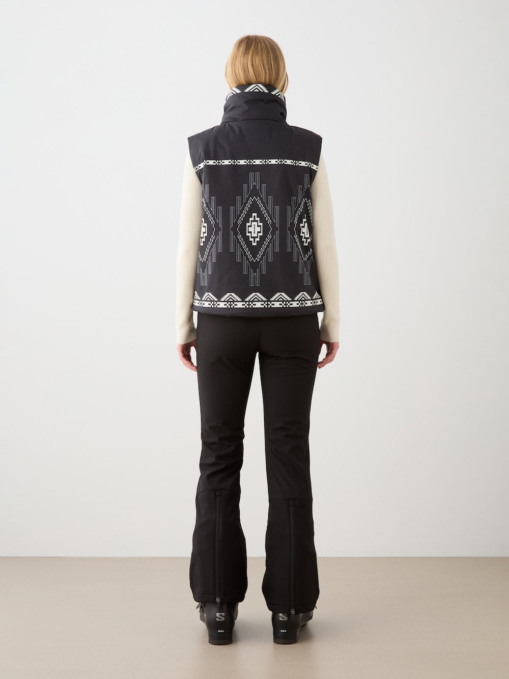 A person stands facing away, wearing the We Norwegians Pioneer Vest for women—a black wool insulated vest with white Indigenous American art-inspired patterns—over a cream sweater and black pants, set against a neutral background.