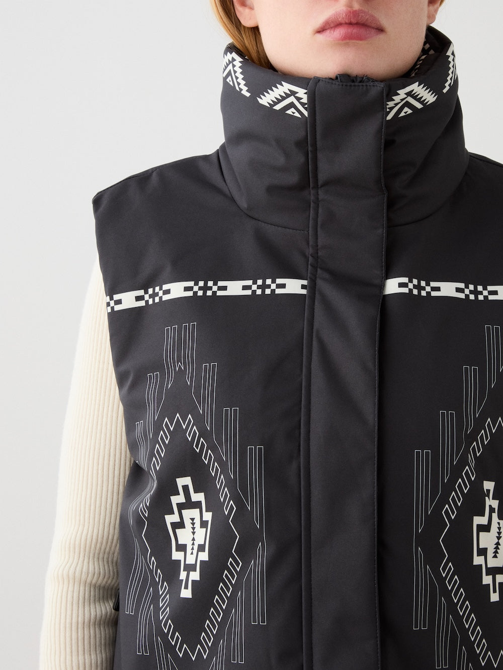 A woman wears the We Norwegians Pioneer Vest—a black digital print vest with white geometric Indigenous American-inspired patterns and high collar—over a cream ribbed sweater, shown from neck to chest.