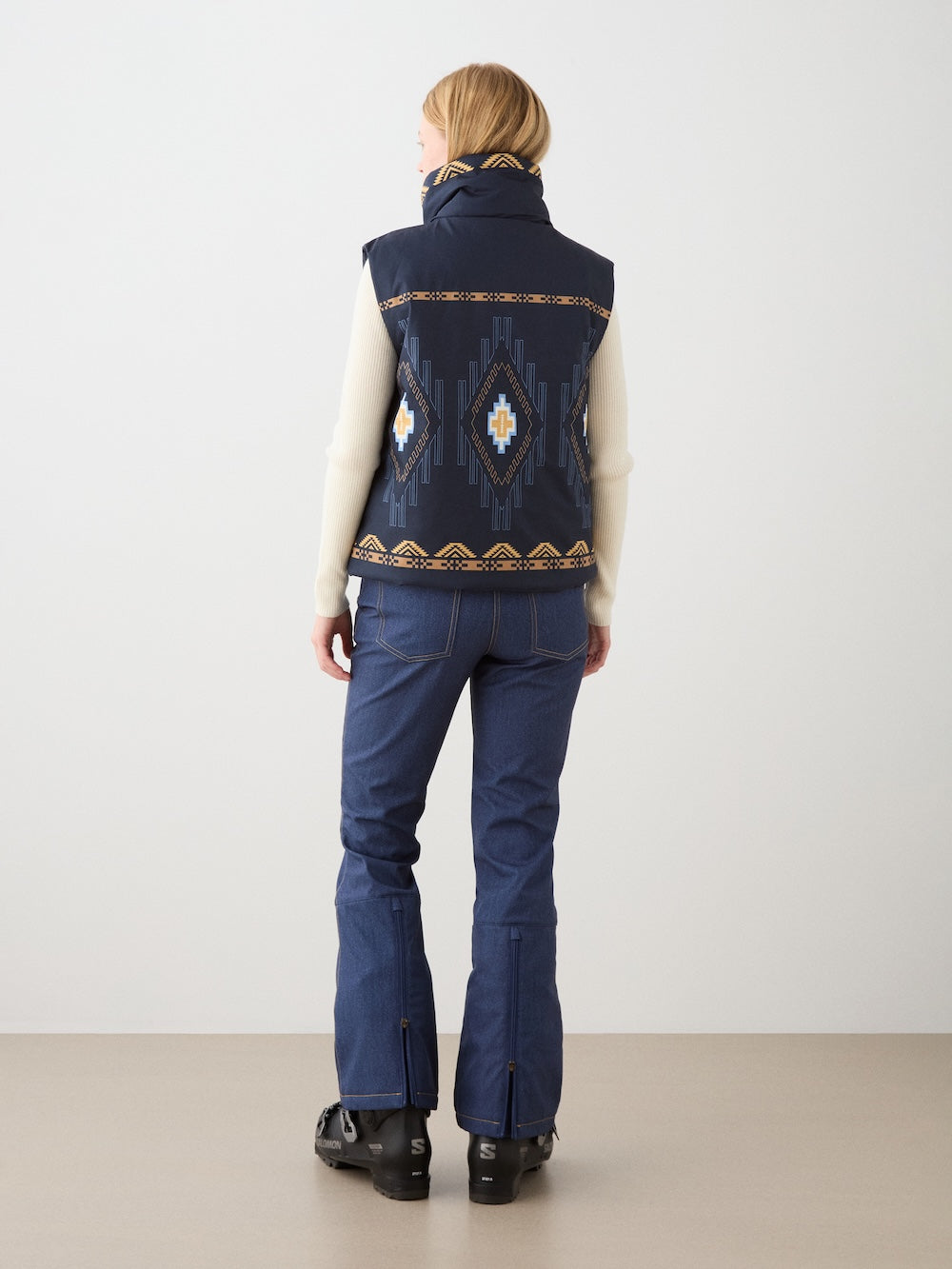 A person faces away, wearing the We Norwegians Pioneer Padded Vest for women in navy with geometric patterns, paired with a cream sweater, blue jeans featuring zipper slits at the back, and black shoes against a plain background.