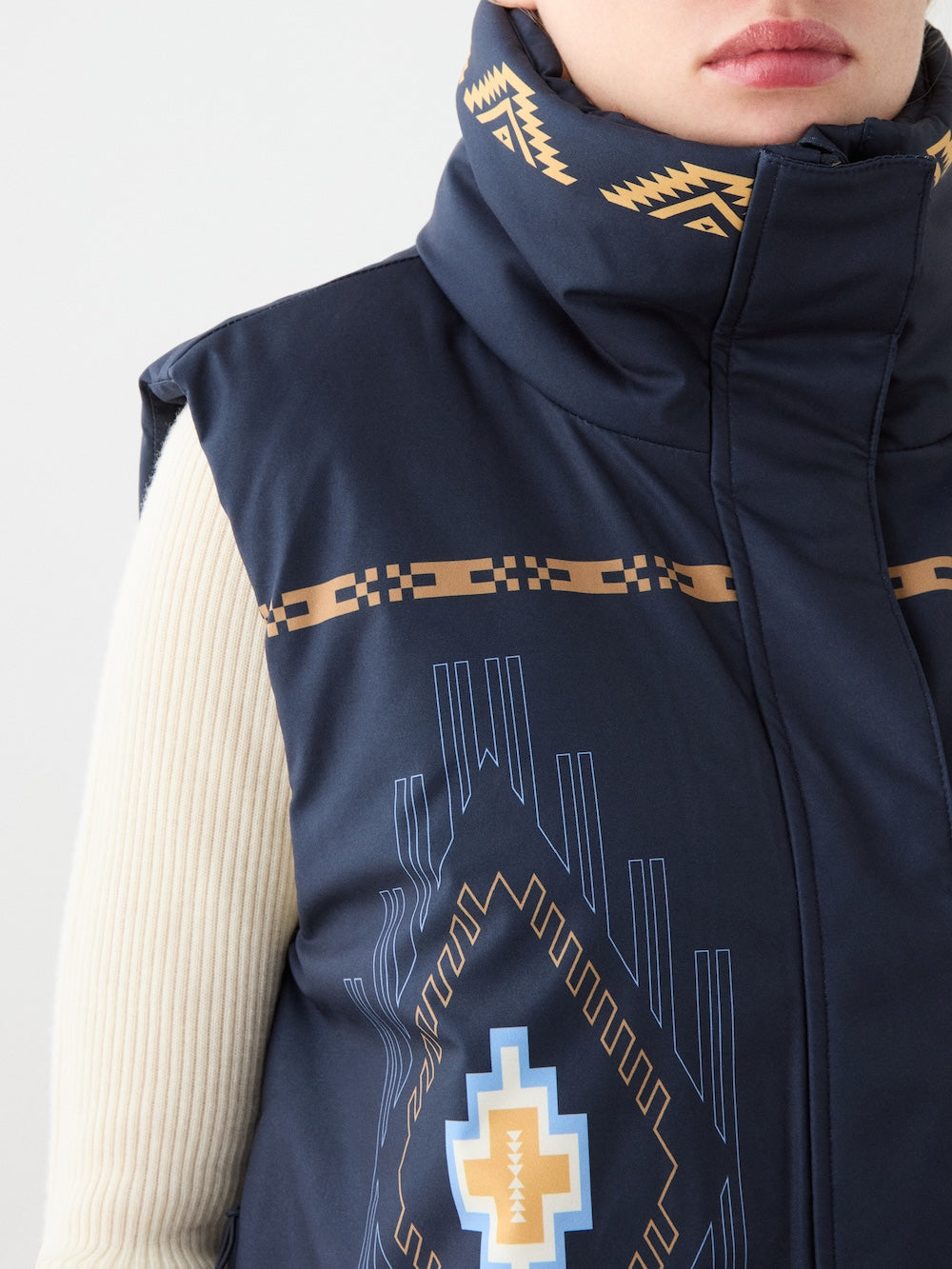 A close-up of a person wearing the We Norwegians Pioneer Vest for women, featuring navy blue digital geometric patterns in beige, white, and blue over a cream ribbed sweater, with a high collar and Indigenous American-inspired designs.
