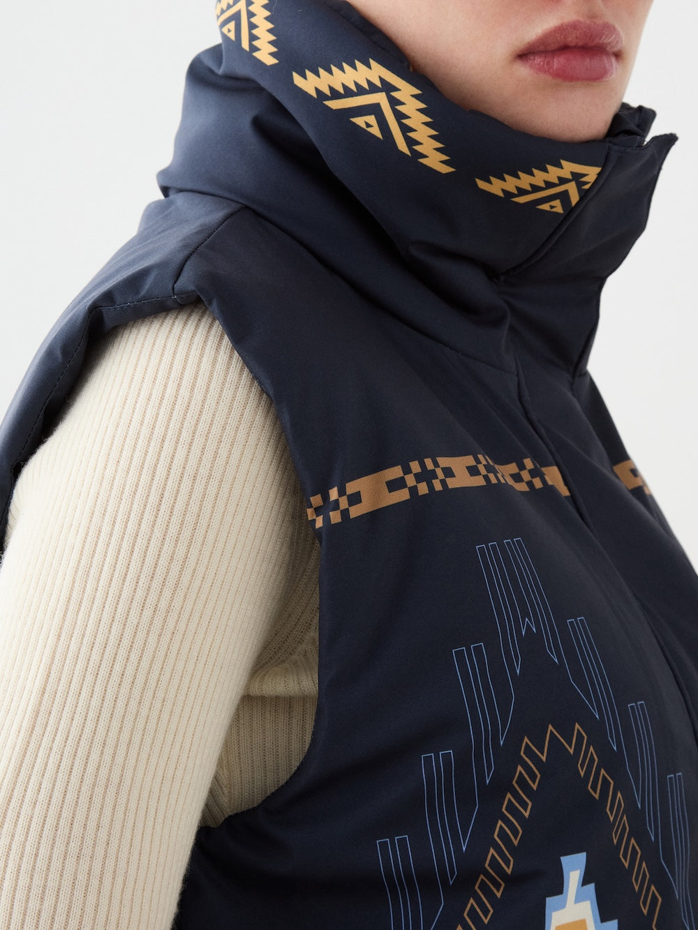 Close-up of a woman wearing the We Norwegians Pioneer Vest, featuring a navy digital print with golden geometric patterns inspired by Indigenous American art, over a cream ribbed turtleneck. The image highlights the vest’s shoulder and collar, with lips partially visible.