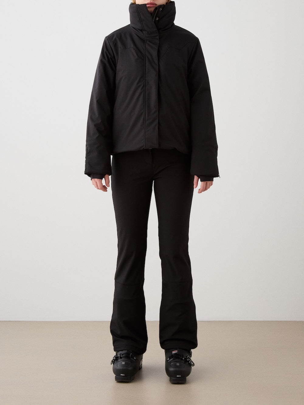 A person wearing the We Norwegians Western Ski Jacket women in black, paired with fitted black pants and boots, stands against a plain light-colored wall. Their face is not visible in the image.