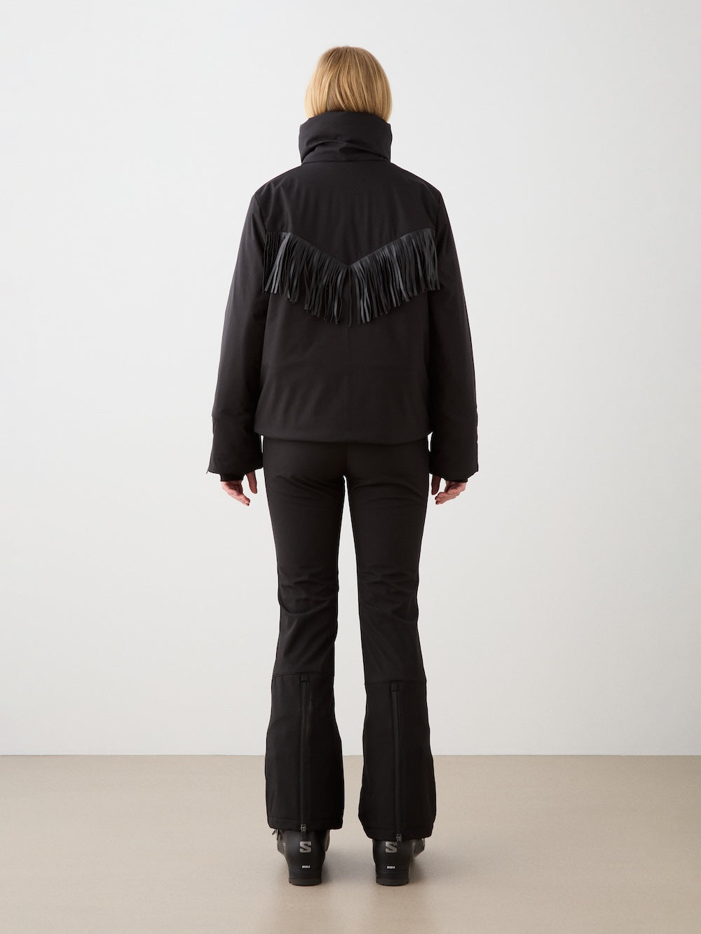 A person with blonde hair, seen from behind, wears the We Norwegians Western Ski Jacket women with fringe detail and matching black pants, standing on a light floor against a plain white wall.
