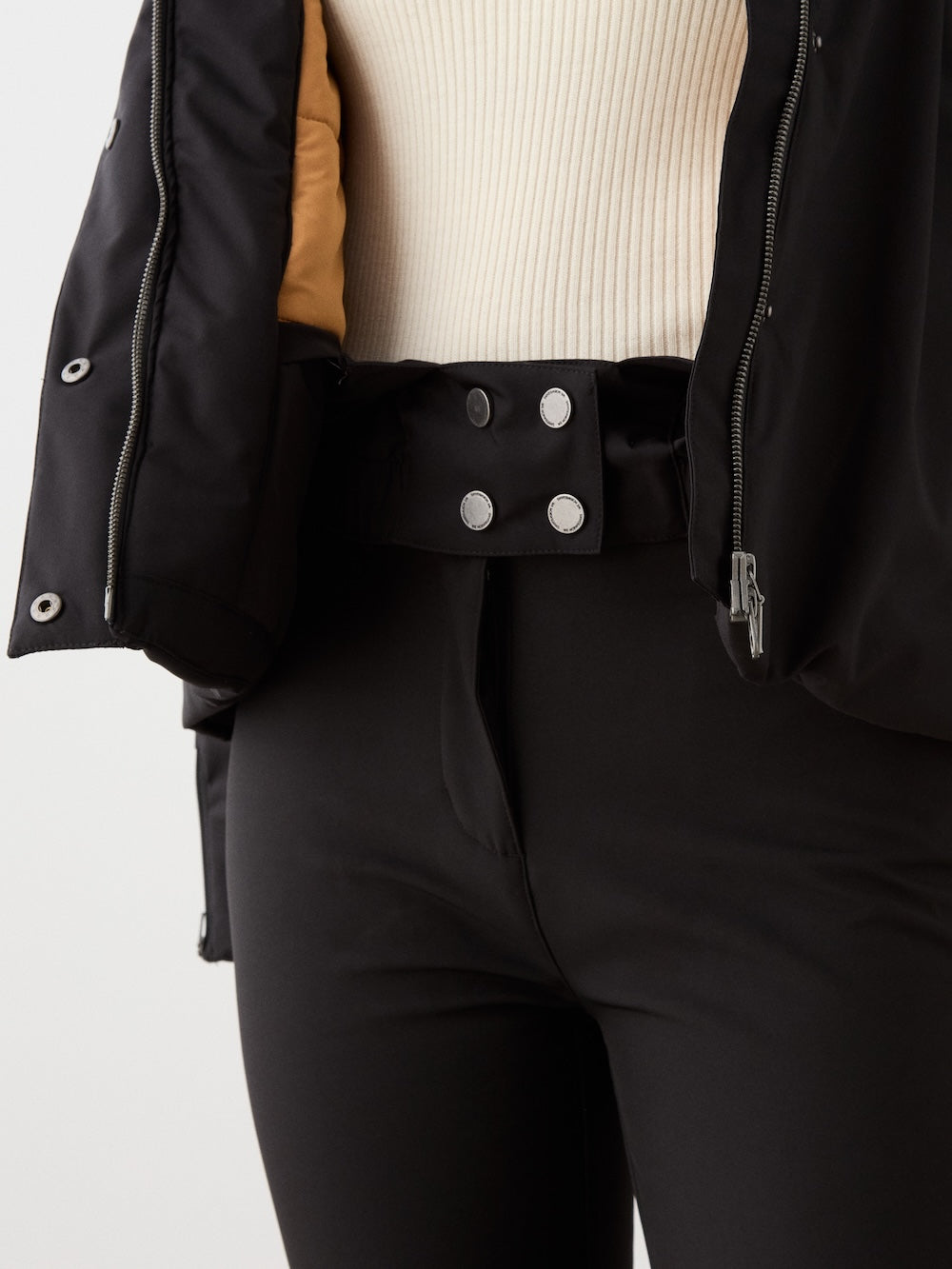A person wears black high-waisted pants with a double-button waistband, a cream ribbed top, and the We Norwegians Western Ski Jacket for women, alpine-inspired with partially exposed inner natural wool padding.