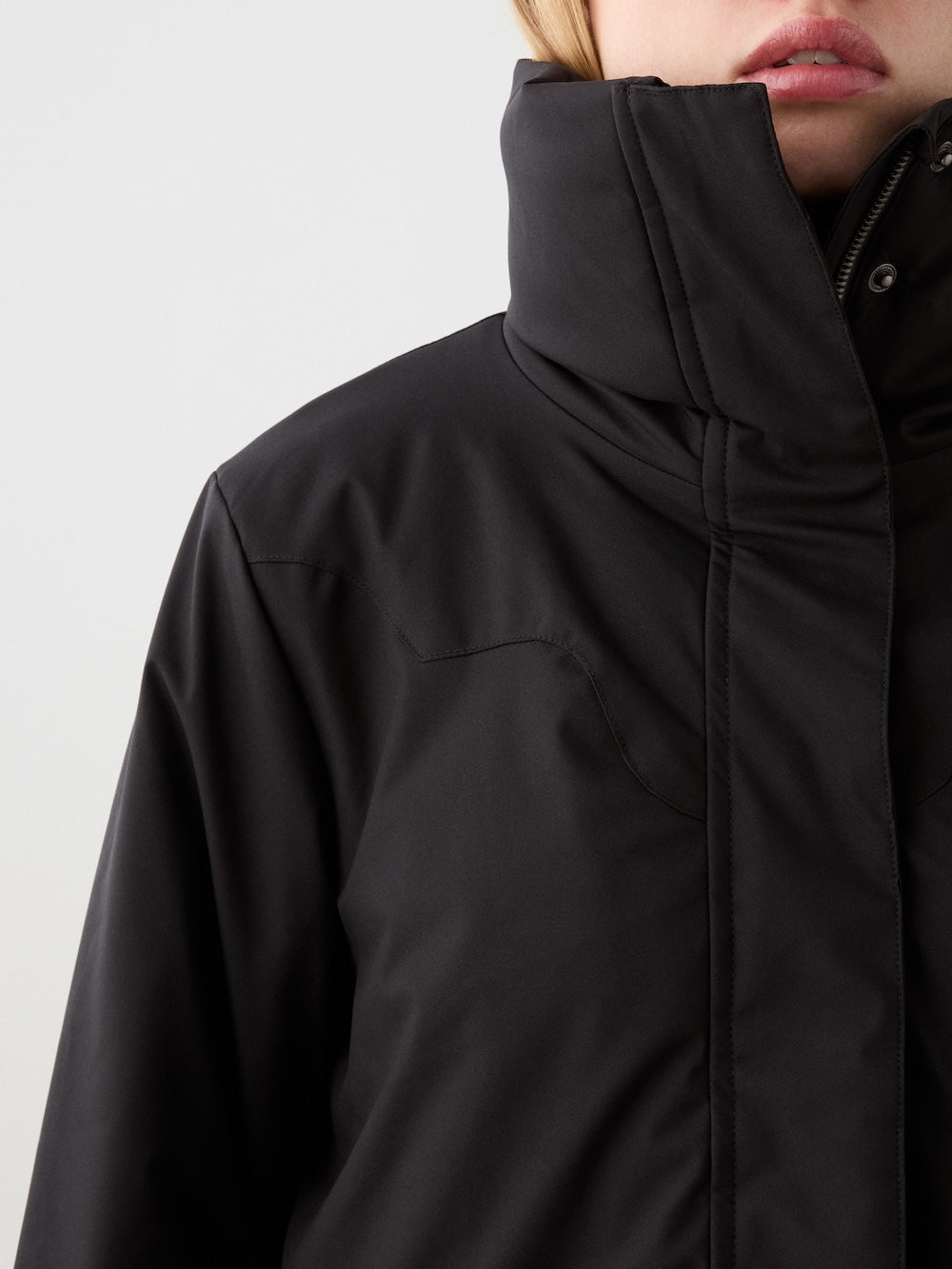 Close-up of a woman wearing the We Norwegians Western Ski Jacket with natural wool padding. The high collar covers part of her light-skinned face, and blond hair is partly visible. Background is plain and light-colored.