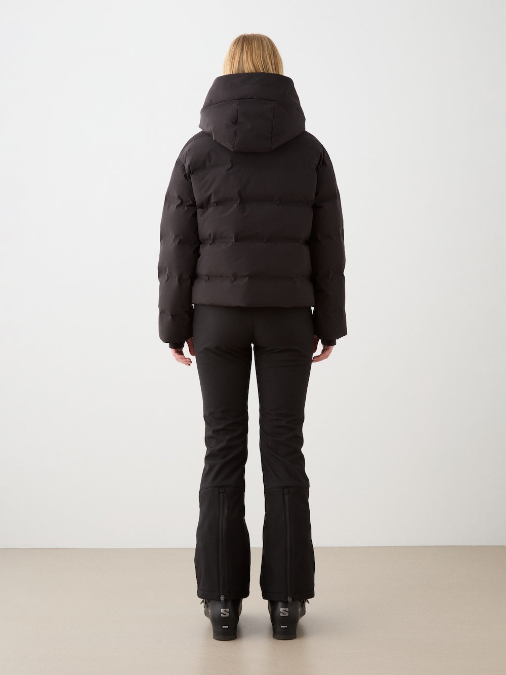 A woman faces away from the camera, wearing the black We Norwegians Aurora Ski Jacket with responsible down, along with fitted pants and black shoes, set against a plain light background.