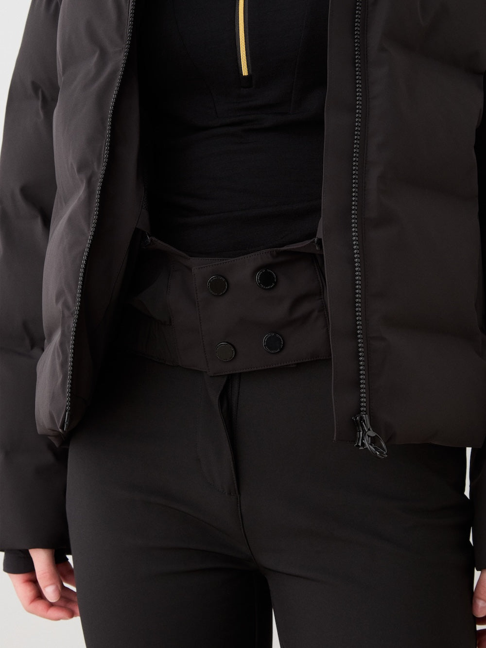 A person wears the We Norwegians Aurora Ski Jacket for women in black, paired with black high-waisted, double-button pants and a black top with a small yellow zipper. The photo is cropped from shoulders to upper thighs.